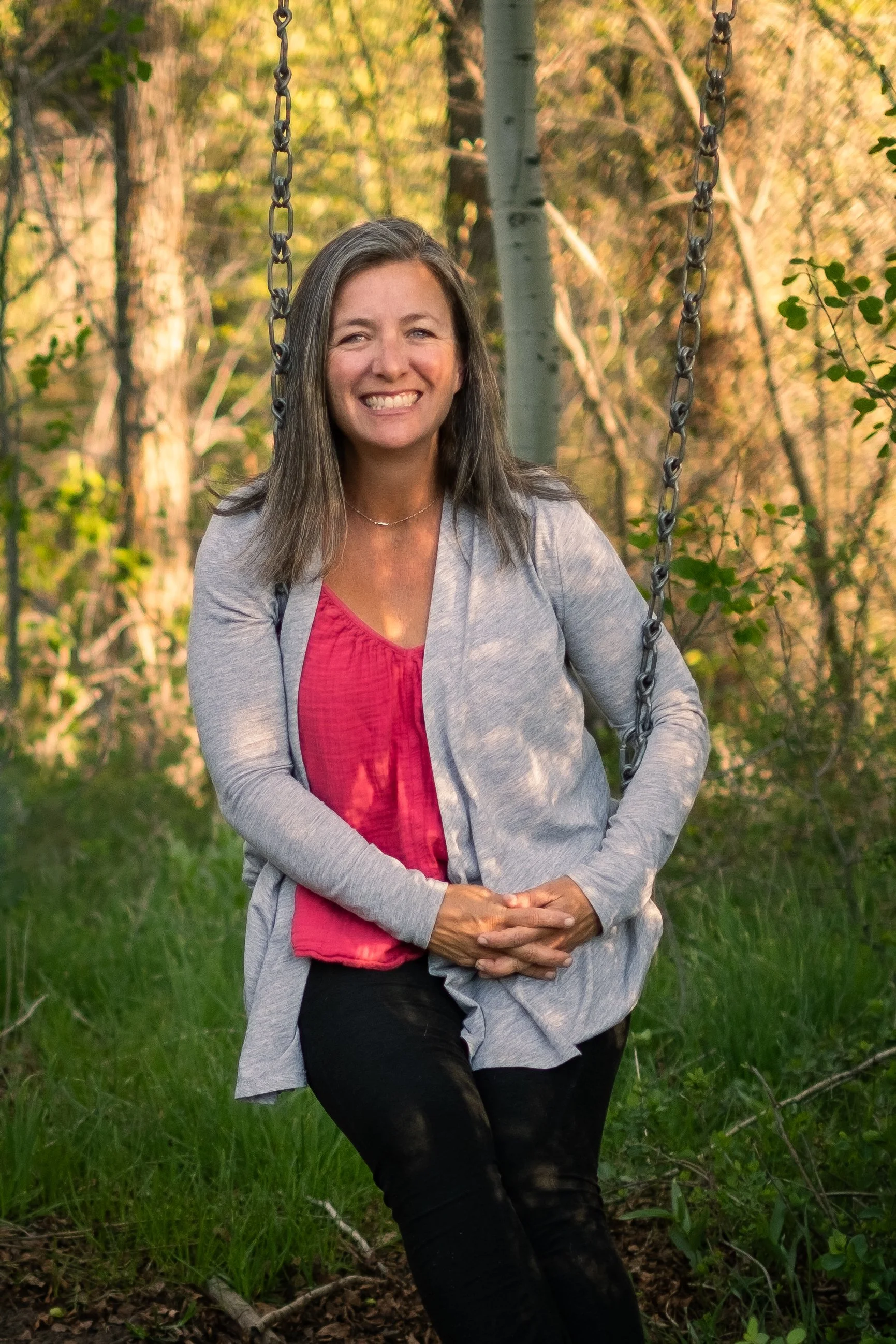 A woman with gray hair smiling on a swing in a forested area with sunlight filtering through trees.