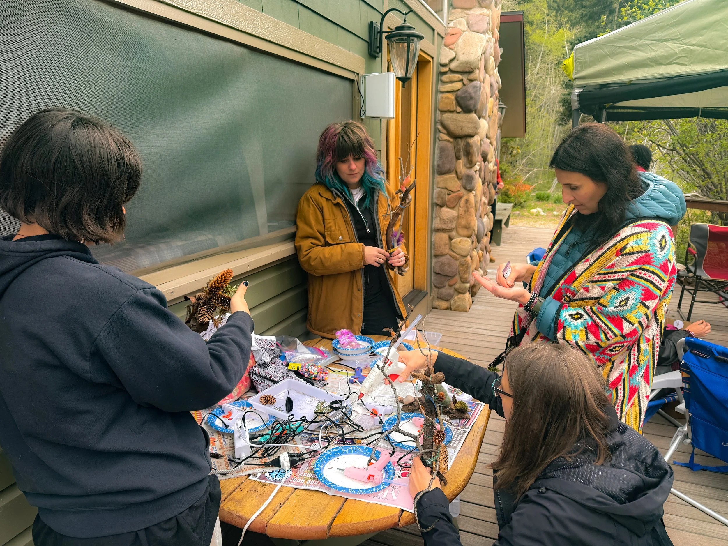 People gathered around a table outdoors, making and displaying arts and crafts such as small tree branches, pinecones, and decorated items, with a house and greenery in the background.