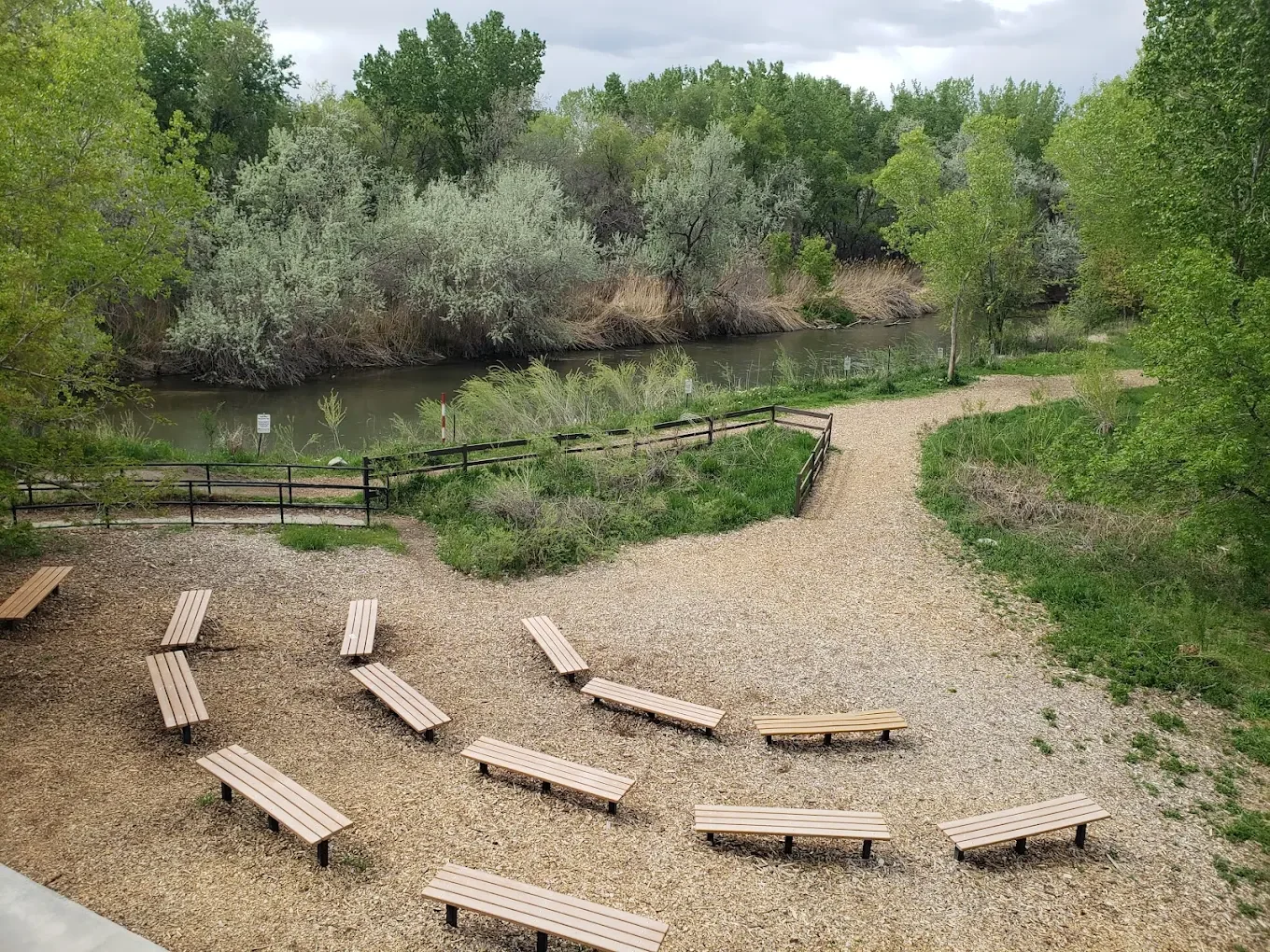A scenic park area with a gravel pathway, wooden benches, a river, and lush green trees.