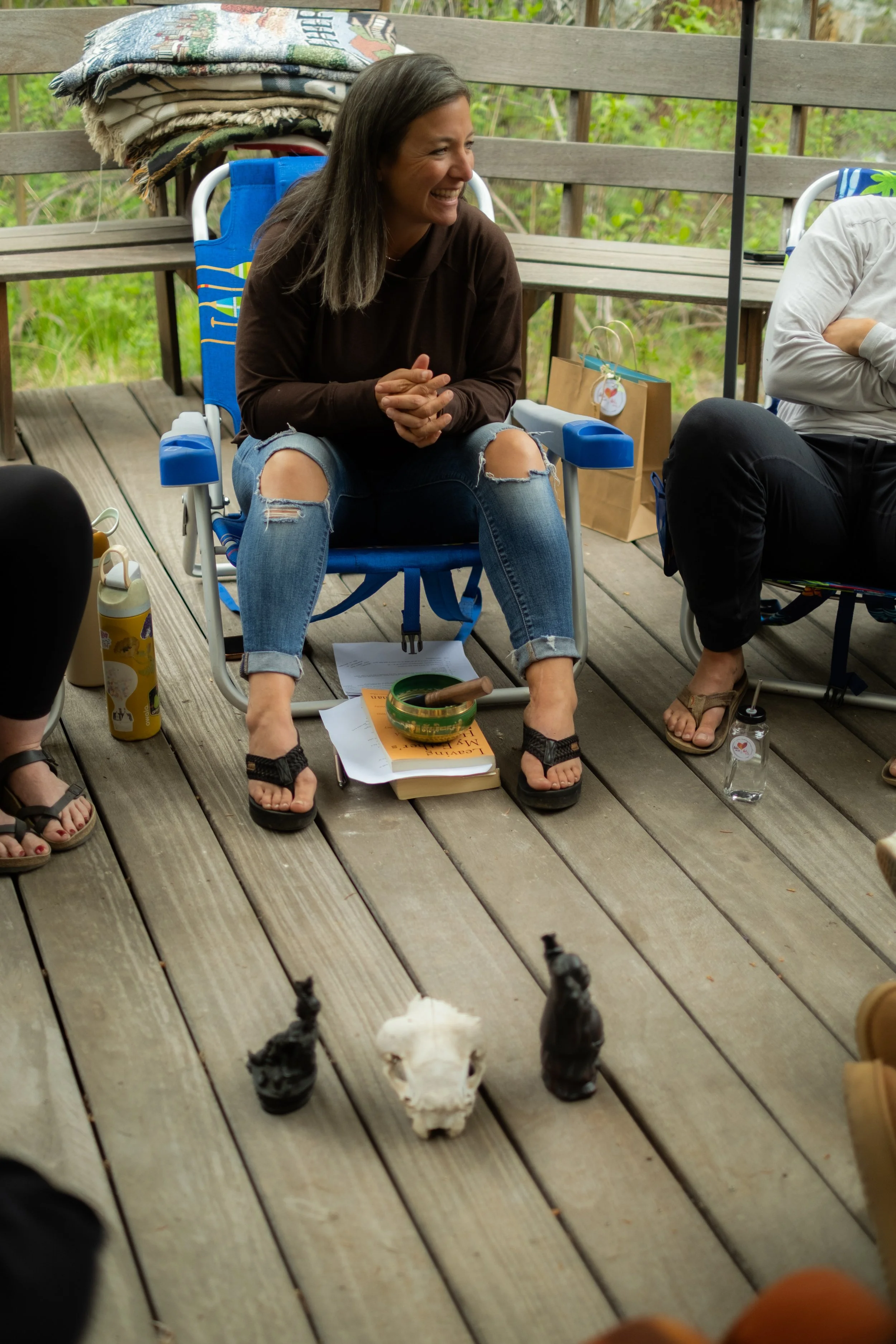 A woman sitting on a folding chair outdoors, smiling and laughing, with a deck and green foliage in the background. There are animals, possibly rabbits or cats, facing her on the wooden deck in front of her.