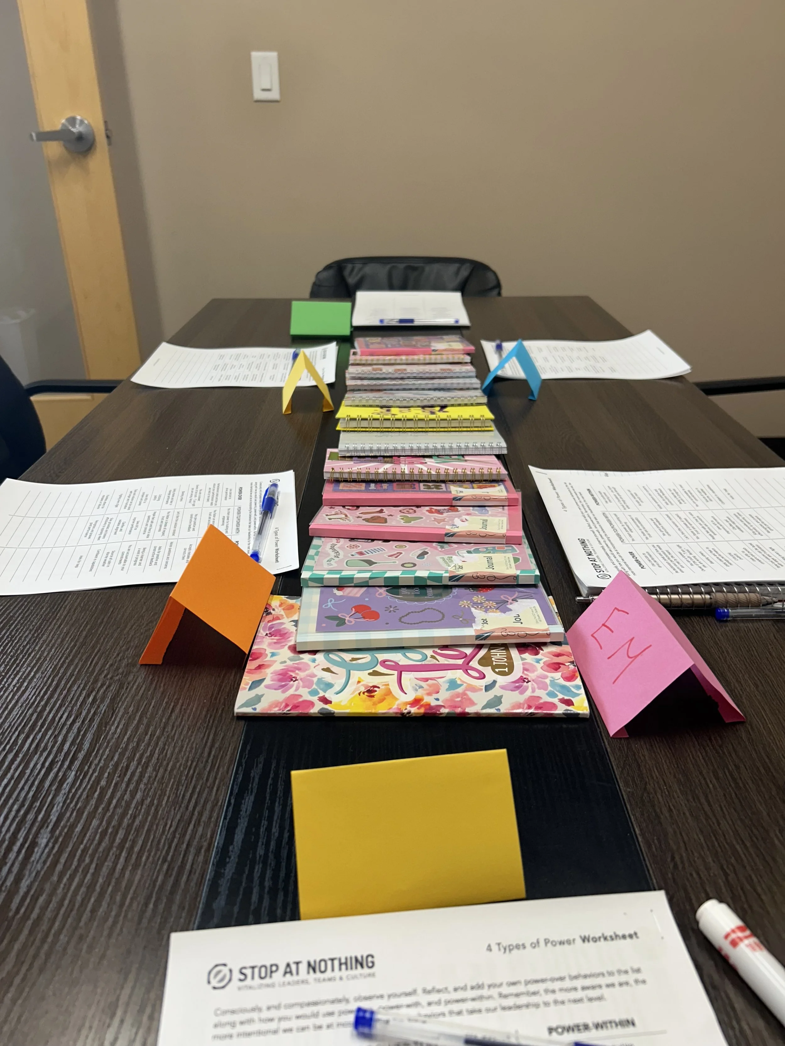 A table with various notebooks, papers, pens, and colorful triangular notes in a meeting or classroom setting.