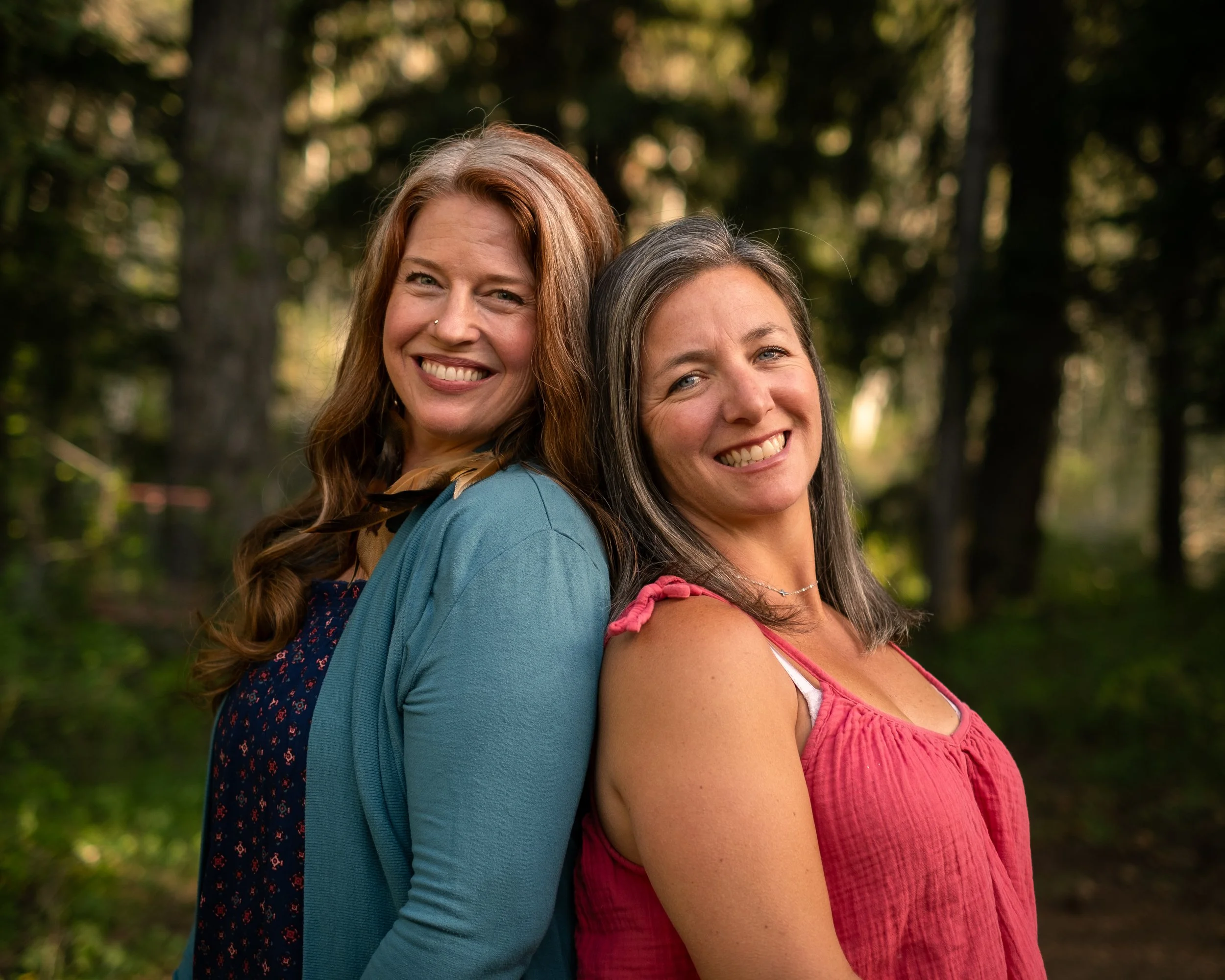Two smiling women standing back to back outdoors in a wooded area.