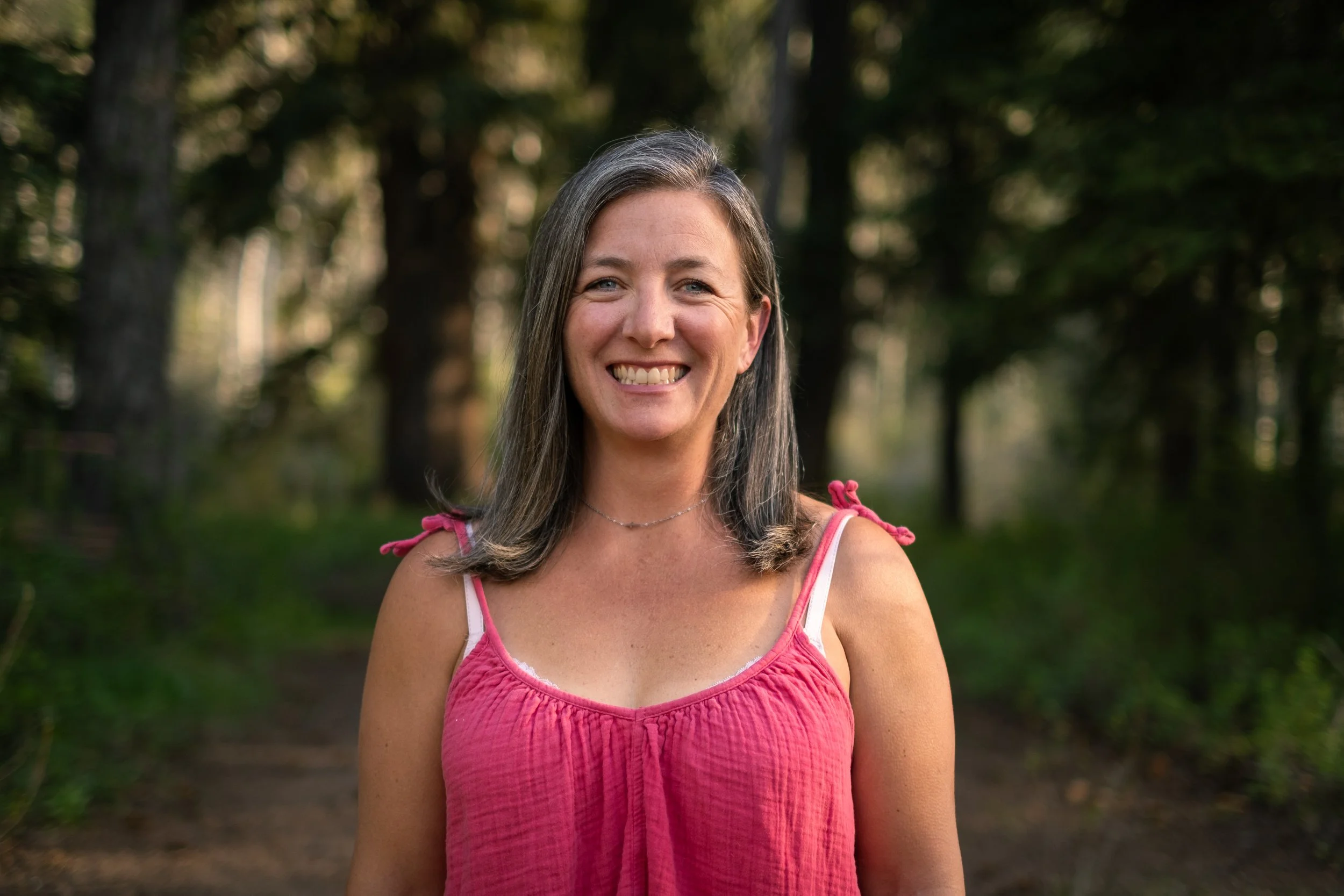 A smiling woman with long gray hair in a pink sleeveless top, standing outdoors with blurred trees and sunlight in the background.