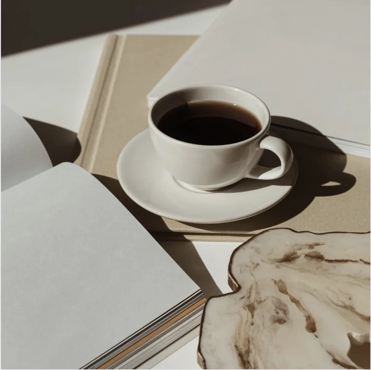 A white cup filled with black coffee on a matching saucer, placed on a beige book, with open blank notebooks around it. A decorative marble tray is partially visible in the foreground.