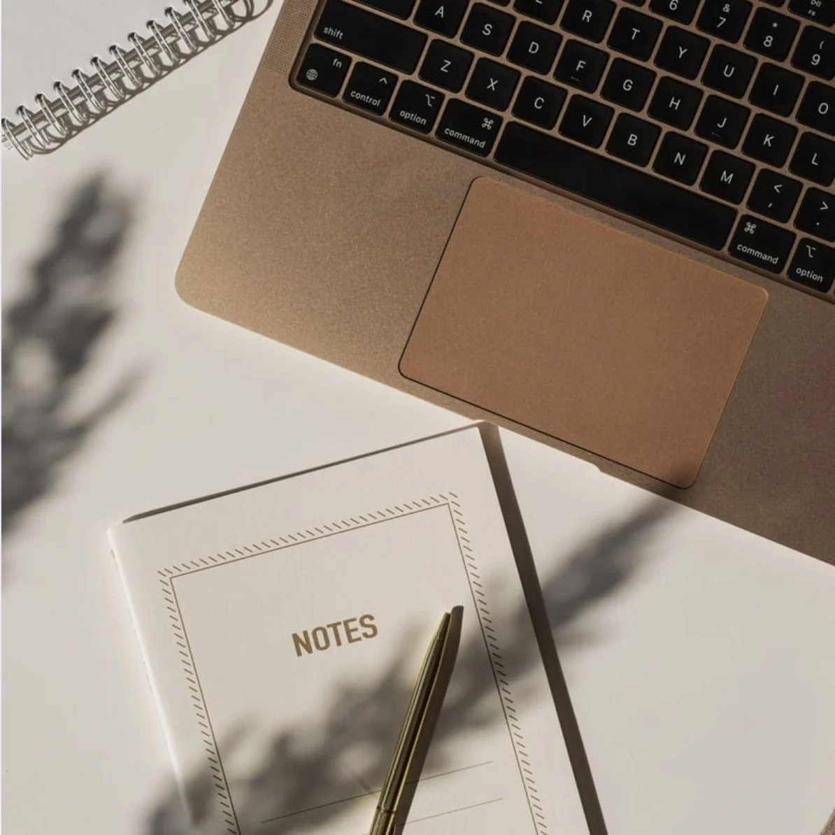 A close-up of a workspace showing the corner of a laptop with a gold finish, a spiral notebook, a gold pen, and a white sheet of paper with the word 'NOTES' printed on it. Shadows and a blurry plant are also visible.