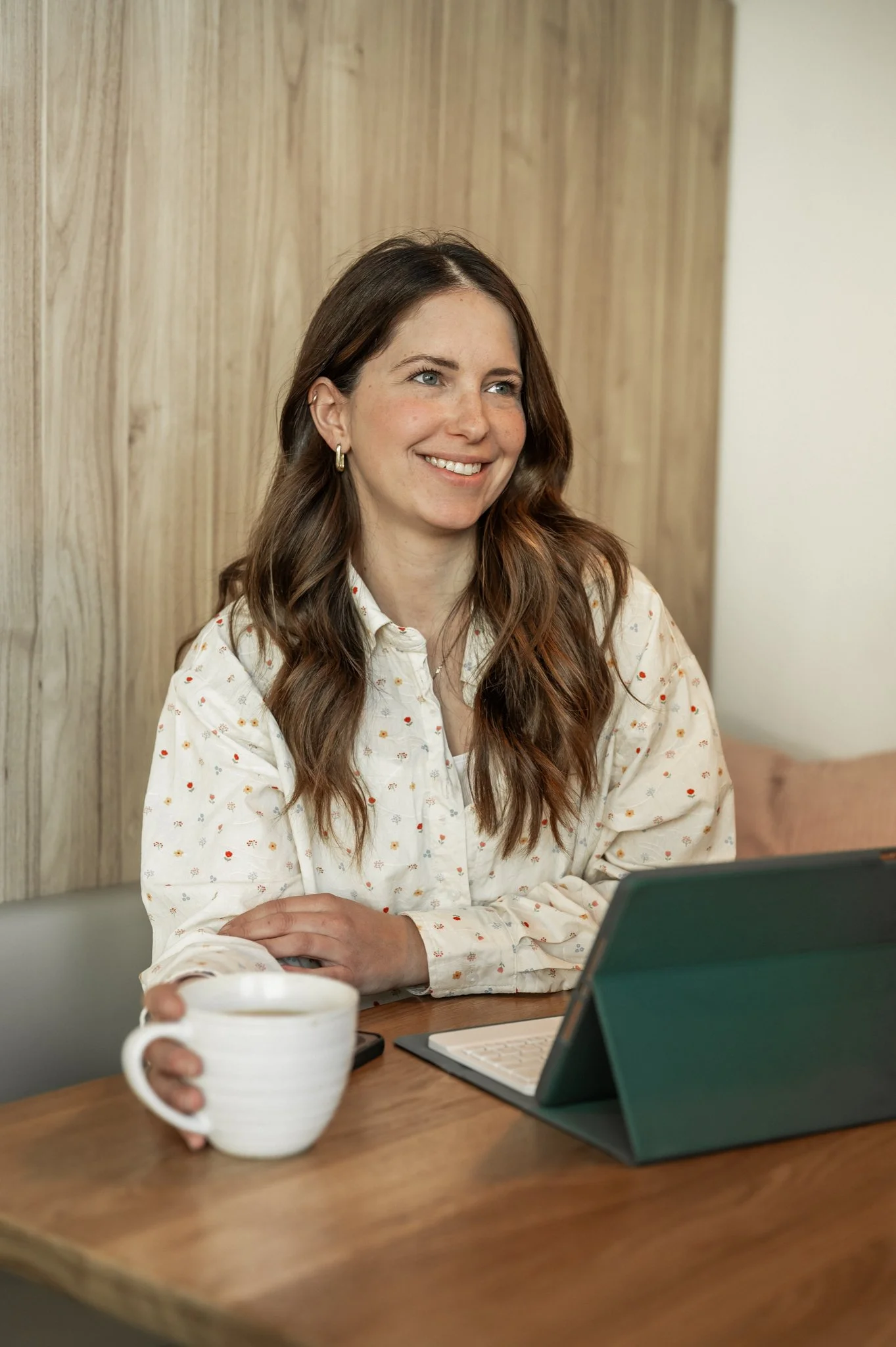 Une femme avec de longs cheveux bruns lisses, souriante, portant une chemise blanche à petits motifs. Elle est assise à une table en bois, tenant une grande tasse blanche, avec un ordinateur portable et une tablette. Le fond comporte un mur en bois clair.