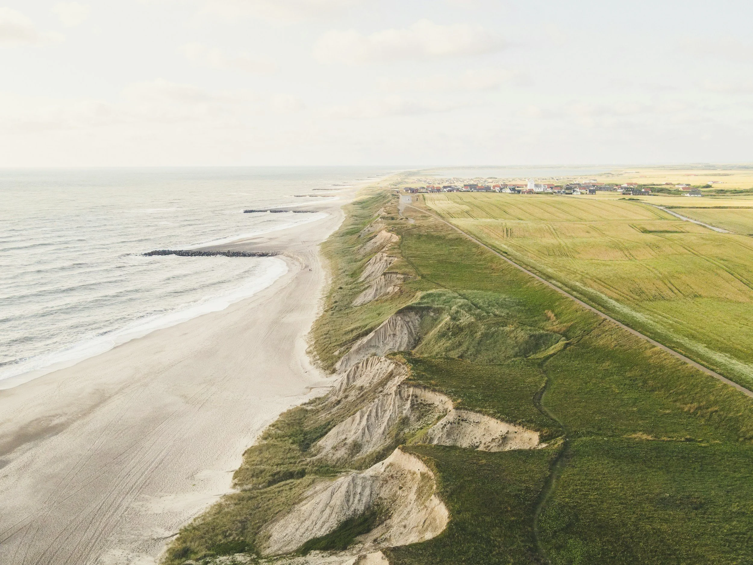 Vues d'une côte avec une plage de sable, des falaises et des champs agricoles. Une petite ville est visible au fond, avec des bâtiments et des champs verts entourant la zone littorale.