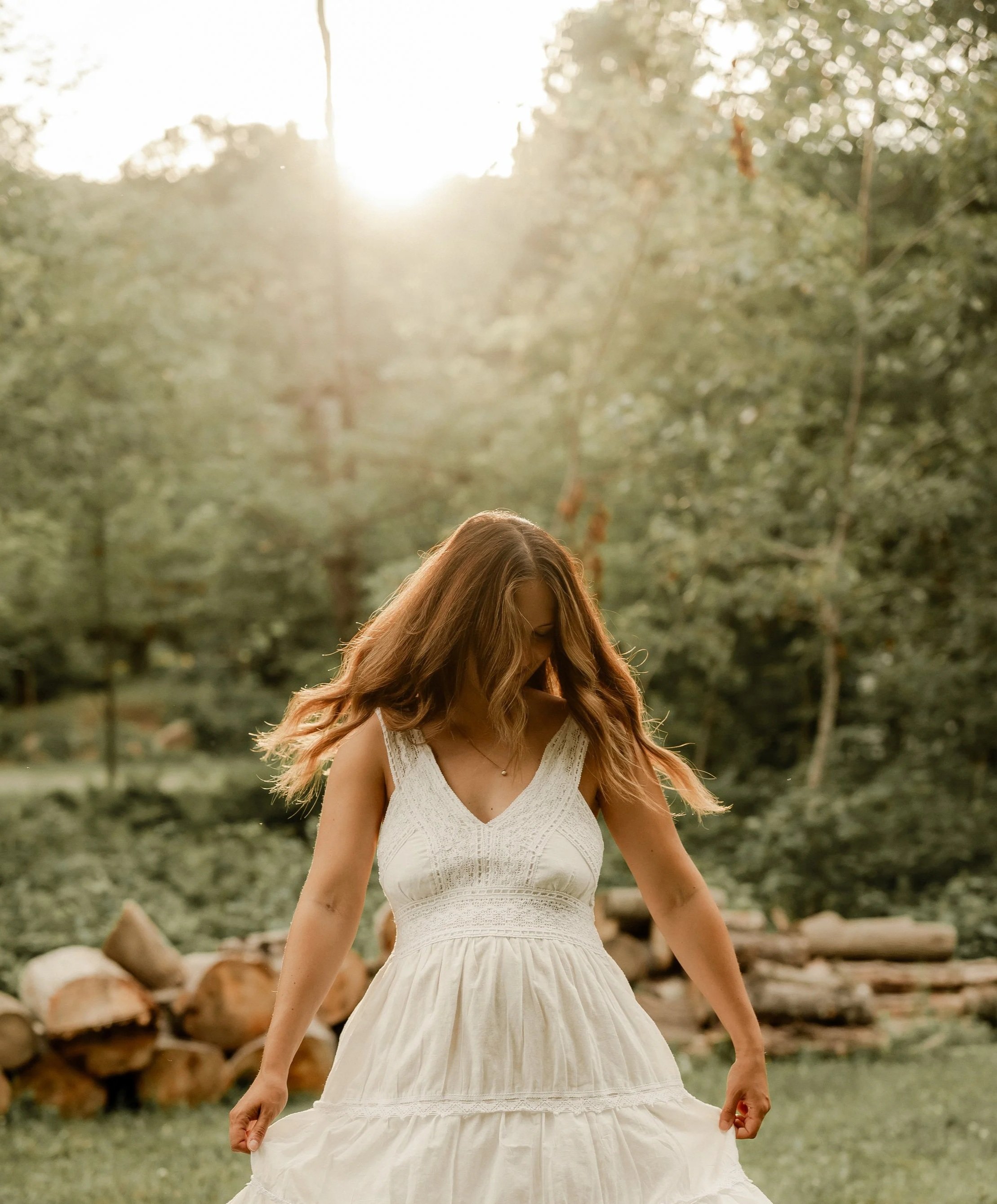 Une femme aux cheveux longs portant une robe blanche, debout dans un environnement naturel avec des arbres et des rochers, la lumière du soleil en arrière-plan.