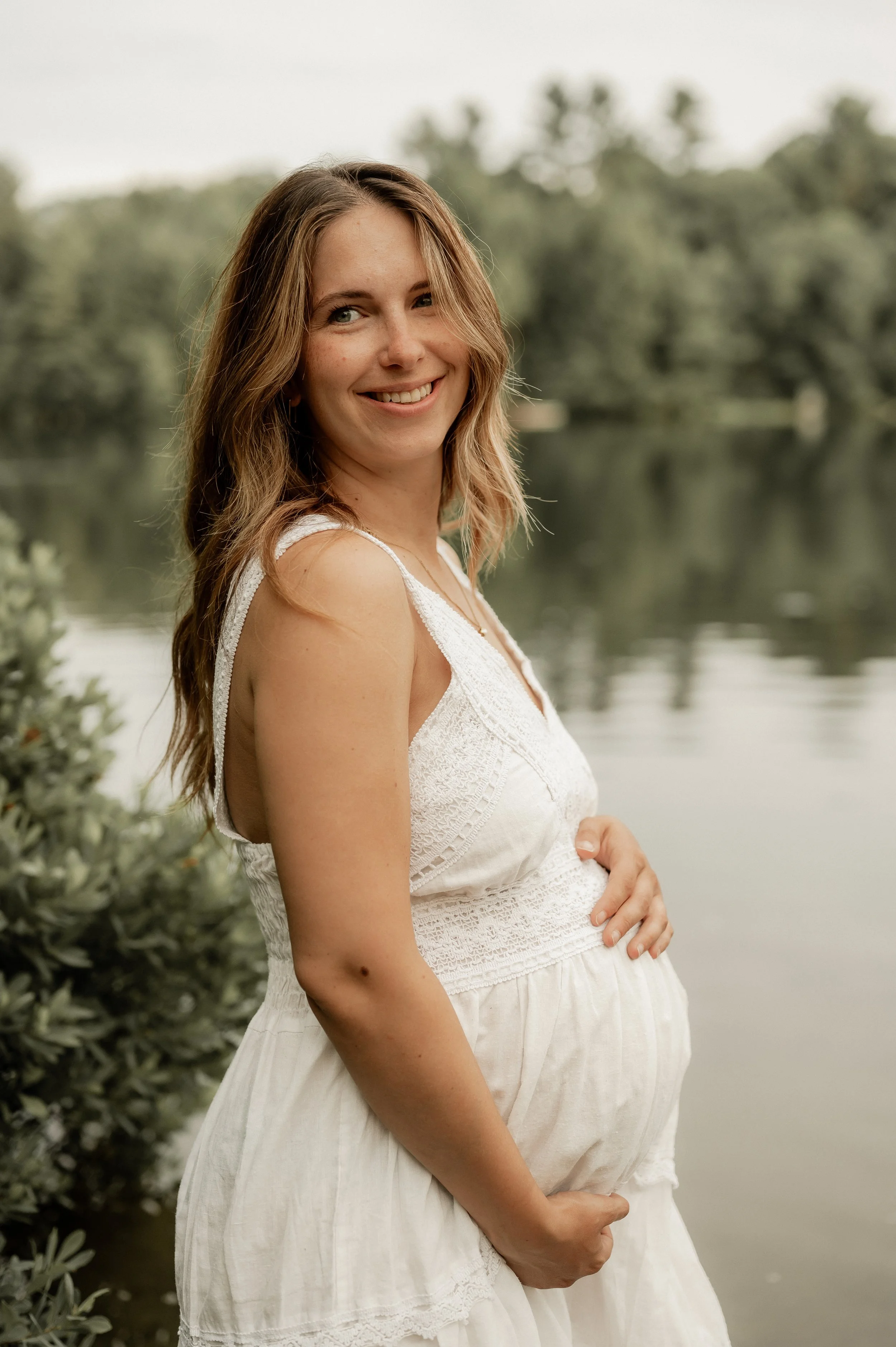 Une femme souriante porte une robe blanche, se tient près d'un lac avec des arbres en arrière-plan.