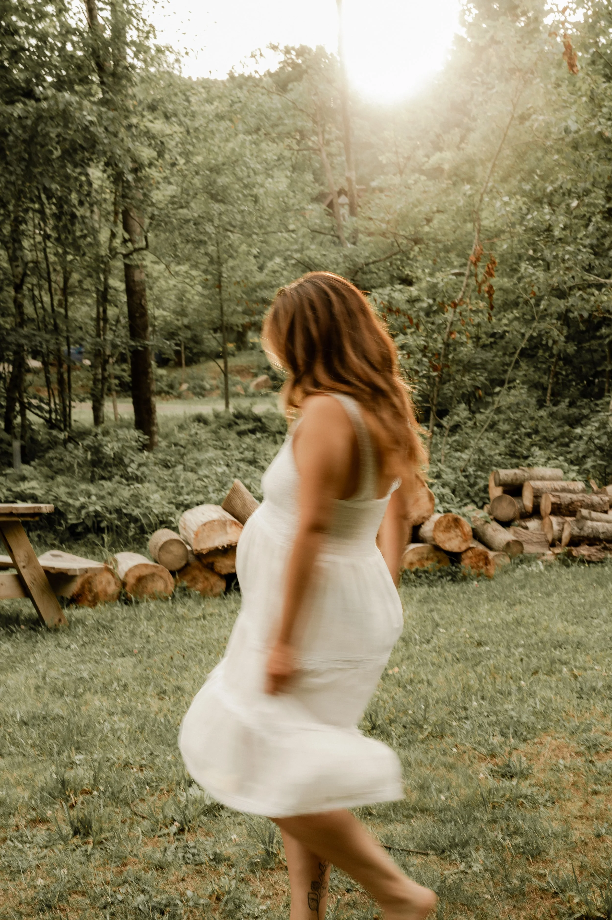 Une femme aux cheveux roux en robe blanche tourne dans un espace extérieur bordé d'arbres pendant la tombée du soleil.