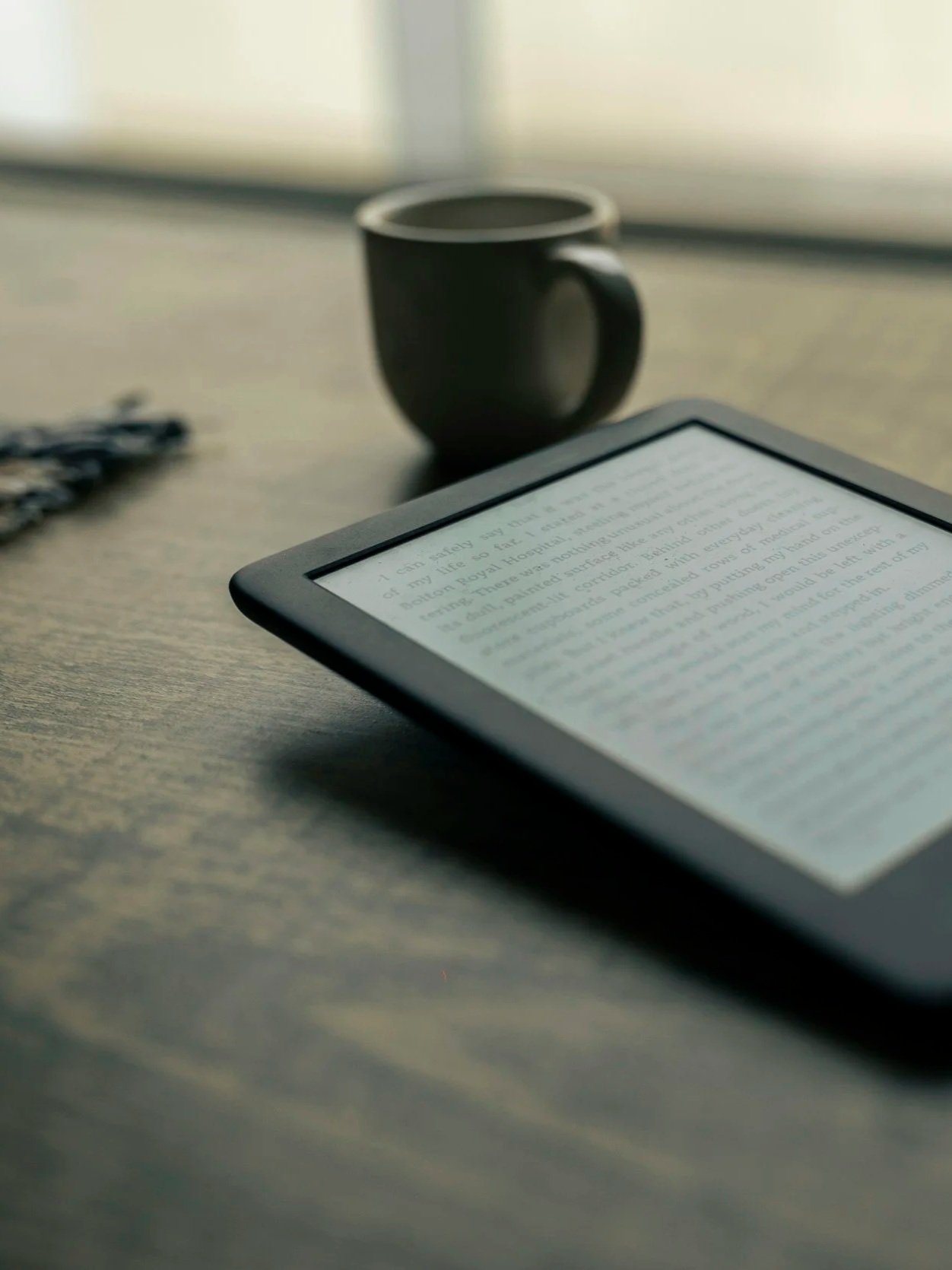 Un livre électronique sur une tablette, une tasse noire en arrière-plan, et une clé sur une surface en bois, dans une pièce bien éclairée.