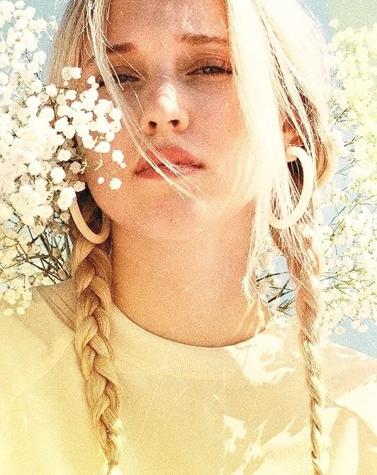 A young woman with blonde hair styled in two braids, wearing large hoop earrings, holding a bunch of small white flowers close to her face, outdoors in sunlight with a blurred background of trees.