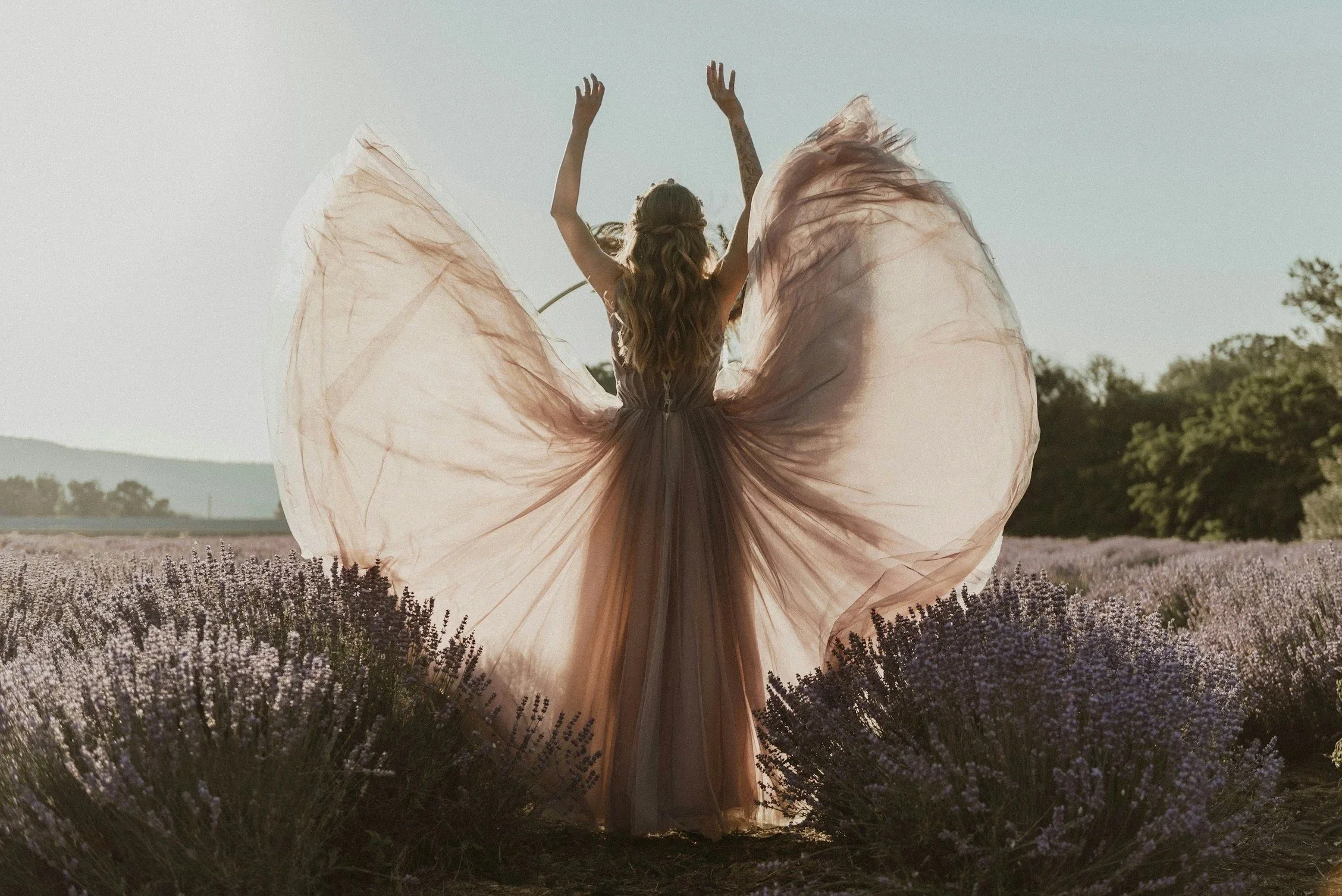 A woman in a flowing, peach-colored dress with large, airy fabric wings stands in a lavender field with trees and hills in the background, arms raised upwards.