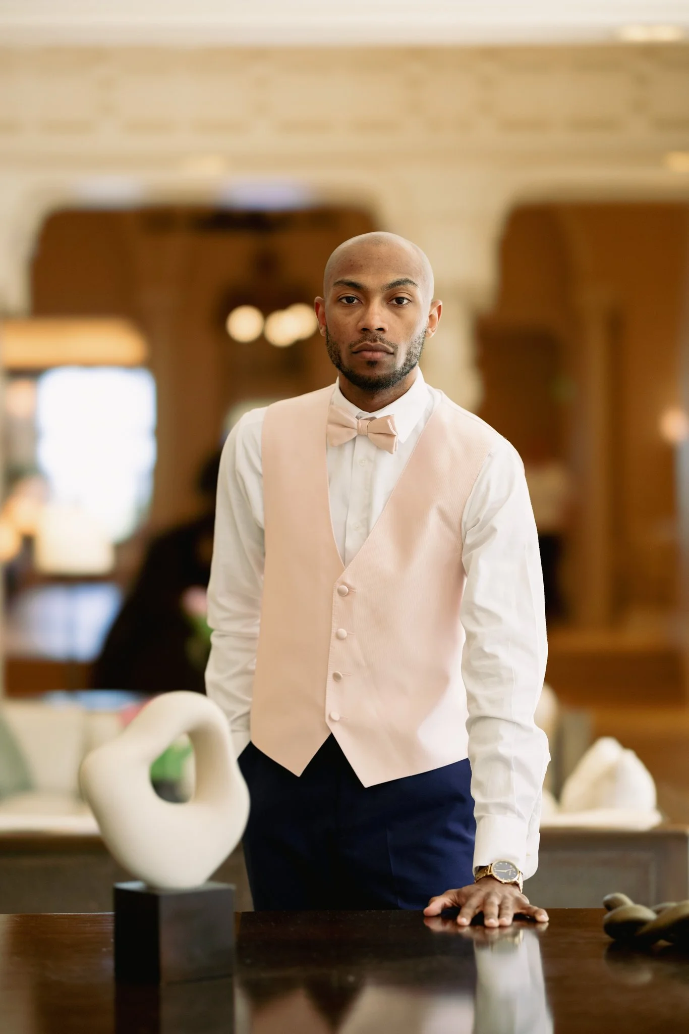 A man with a shaved head and beard wearing a white shirt, beige vest, and bow tie standing with one hand on a table in an elegant room.