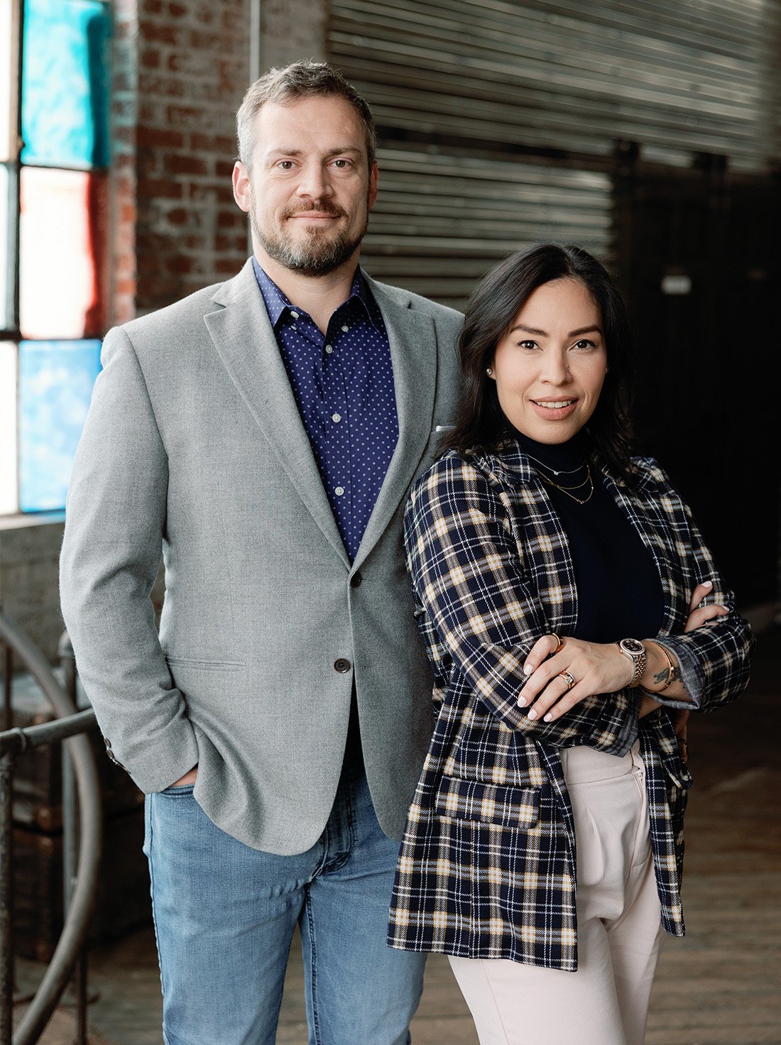 A man and a woman standing indoors with brick walls and a large window in the background. The man has a short beard, is wearing a gray blazer over a blue polka dot shirt, and has his hands in his pockets. The woman has shoulder-length black hair, is wearing a plaid blazer over a black top, and has her arms crossed. The woman is smiling and they are both looking at the camera.