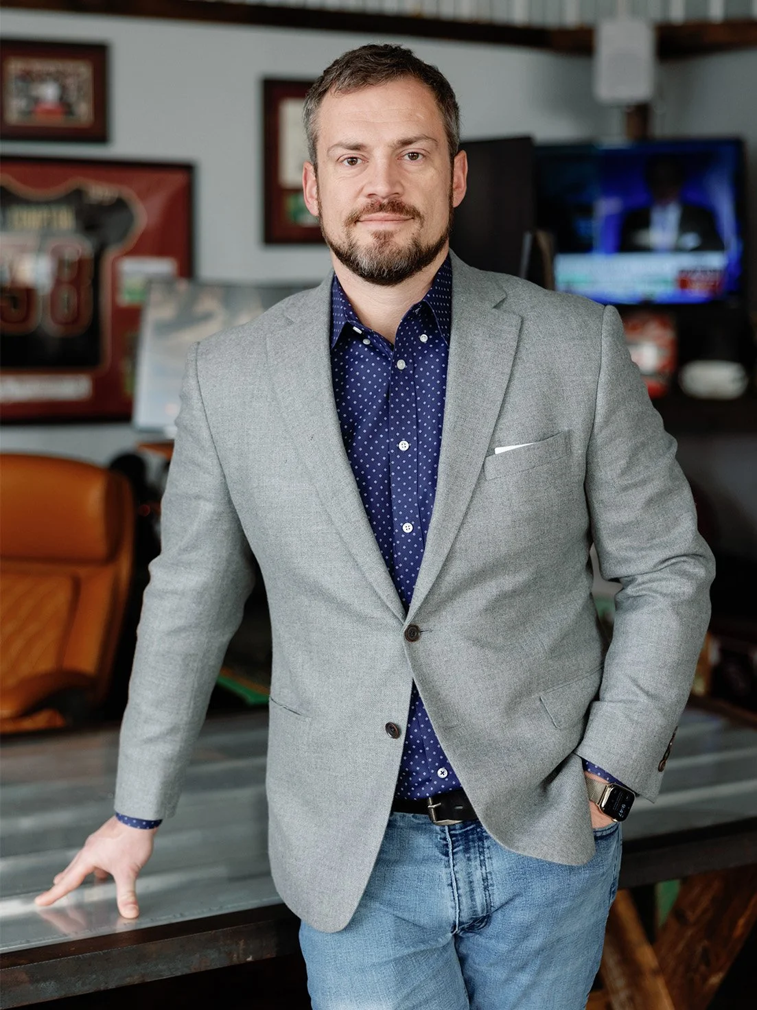 A man in a gray blazer and blue polka dot shirt standing with one arm resting on a wooden table in a room with framed pictures and a television in the background.