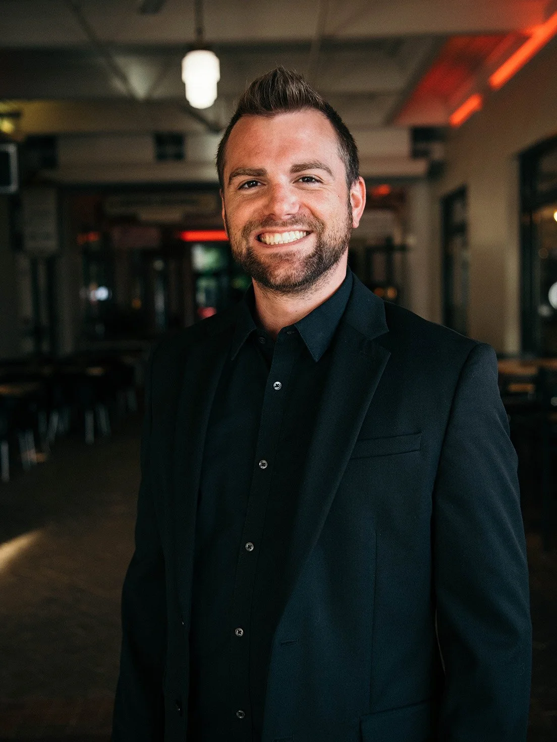 Smiling man with beard in black suit and shirt standing in a dimly lit room with tables and chairs.