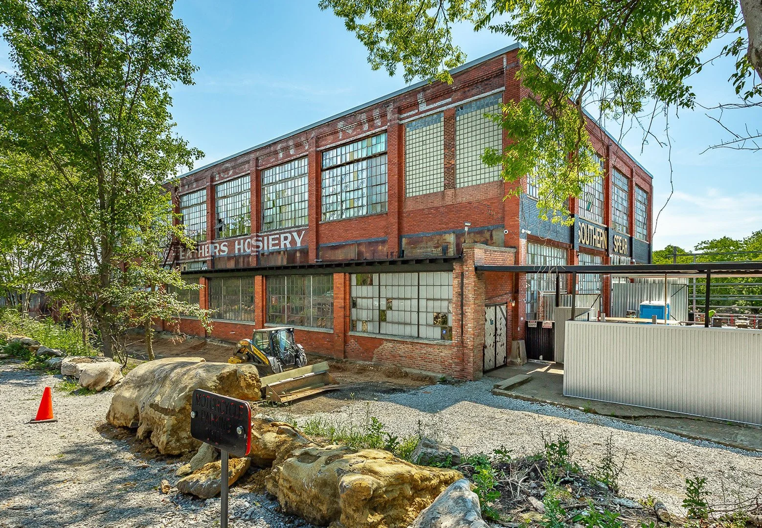 An old, abandoned brick building with large industrial windows and faded signs that read 'Wethers Hosiery' and 'Southern Sewing'. There is a small construction vehicle and rocks in the foreground, with some trees and a gravel pathway leading around the building.