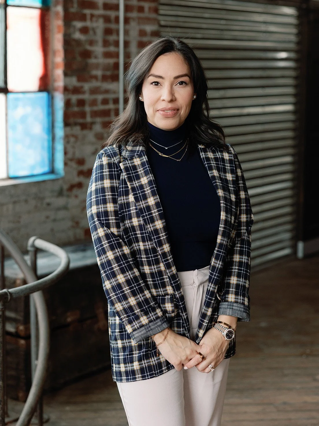 A woman with medium-length dark hair and light skin posing indoors in front of a brick wall and window, wearing a plaid blazer, a black turtleneck, light-colored pants, and jewelry.