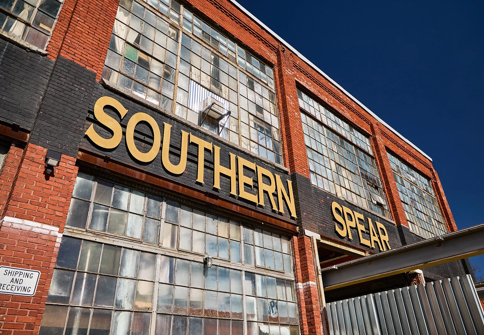 Exterior of a red brick industrial building with large multi-pane windows and a black horizontal sign with large gold letters that say 'SOUTHERN SPEAR.'