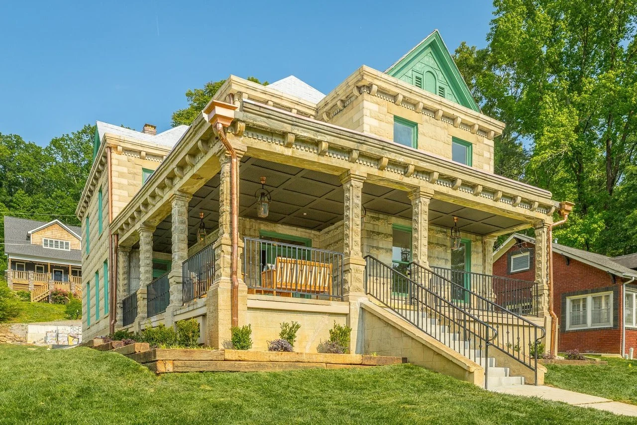 A large stone house with a green roof and a wrap-around porch, surrounded by greenery and trees, with a clear blue sky overhead.