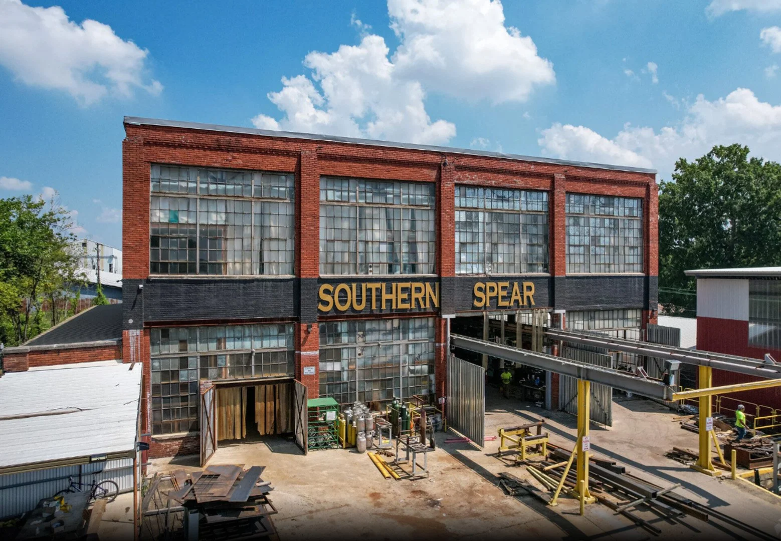 A multi-story brick building with large glass windows and a black sign with gold letters reading 'SOUTHERN SPEAR'. There are construction materials and equipment in the foreground, suggesting ongoing renovation or construction work.