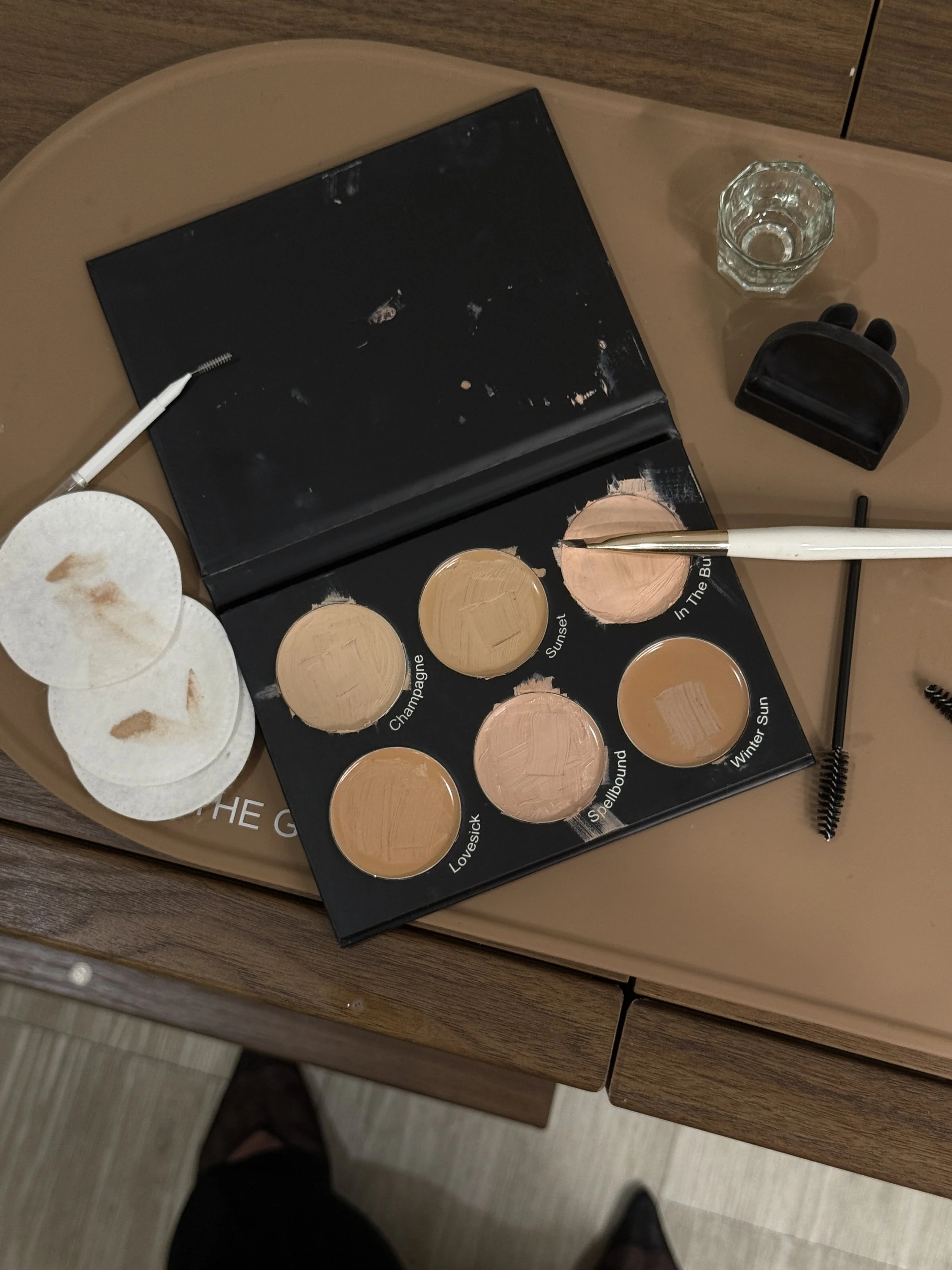 Makeup palette with six shades labeled with names like Champagne, Sunset, Lovesick, Spellbound, In The Bum, and Winter Sun, placed on a brown table along with cotton pads, a glass of water, and eyebrow brushes.