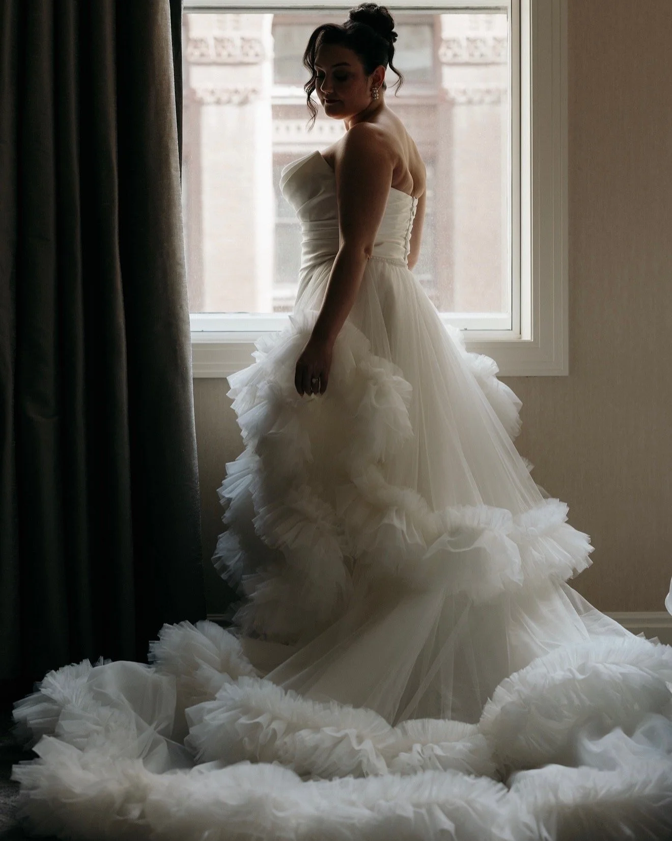 A woman in a wedding dress standing by a window with curtains, with her head slightly tilted down and her silhouette softly lit from outside.