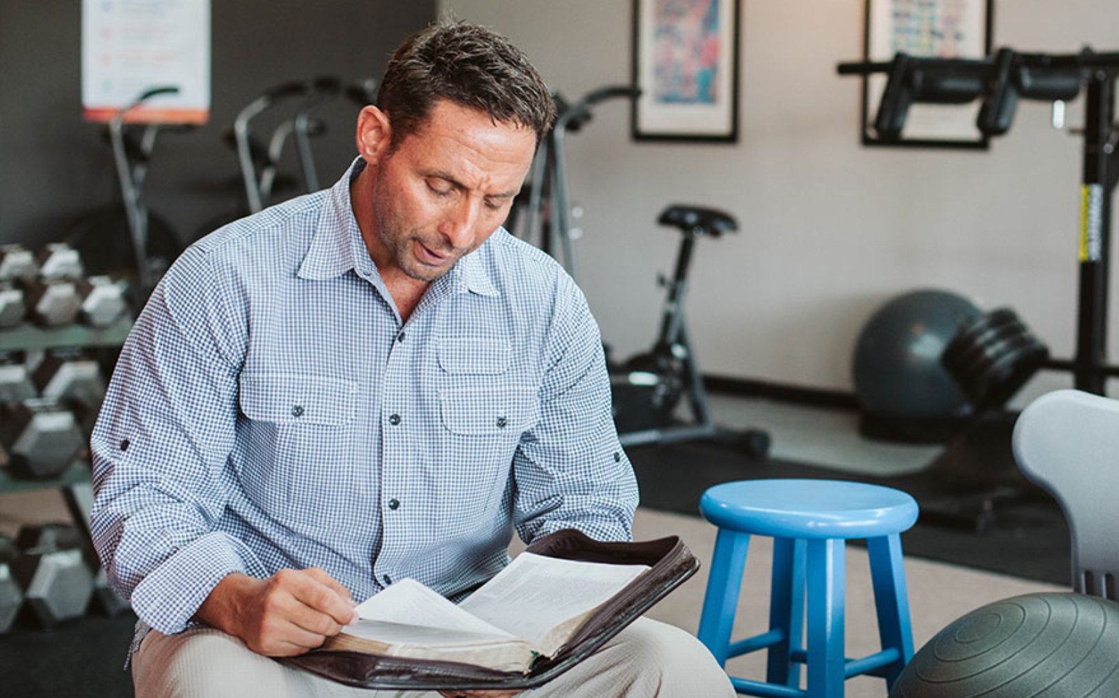 A man sitting in a gym, reading a book, with workout equipment including dumbbells, a exercise bike, a medicine ball, and kettlebells in the background.