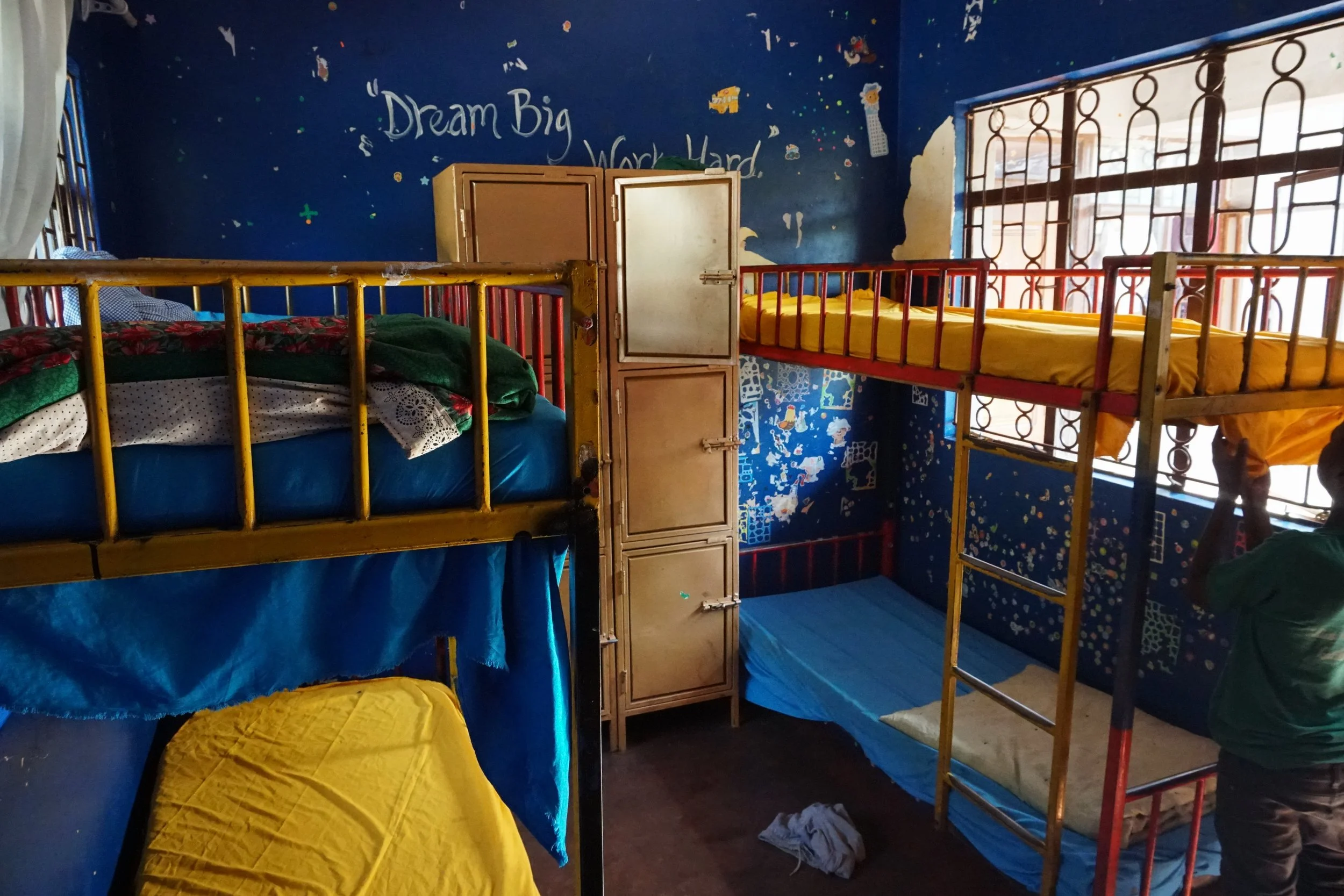 A small room with three bunk beds, a storage cabinet, and a window. The walls are painted blue with colorful decorations and a motivational quote that says 'Dream Big Work Hard.' One person is partially visible in the corner.