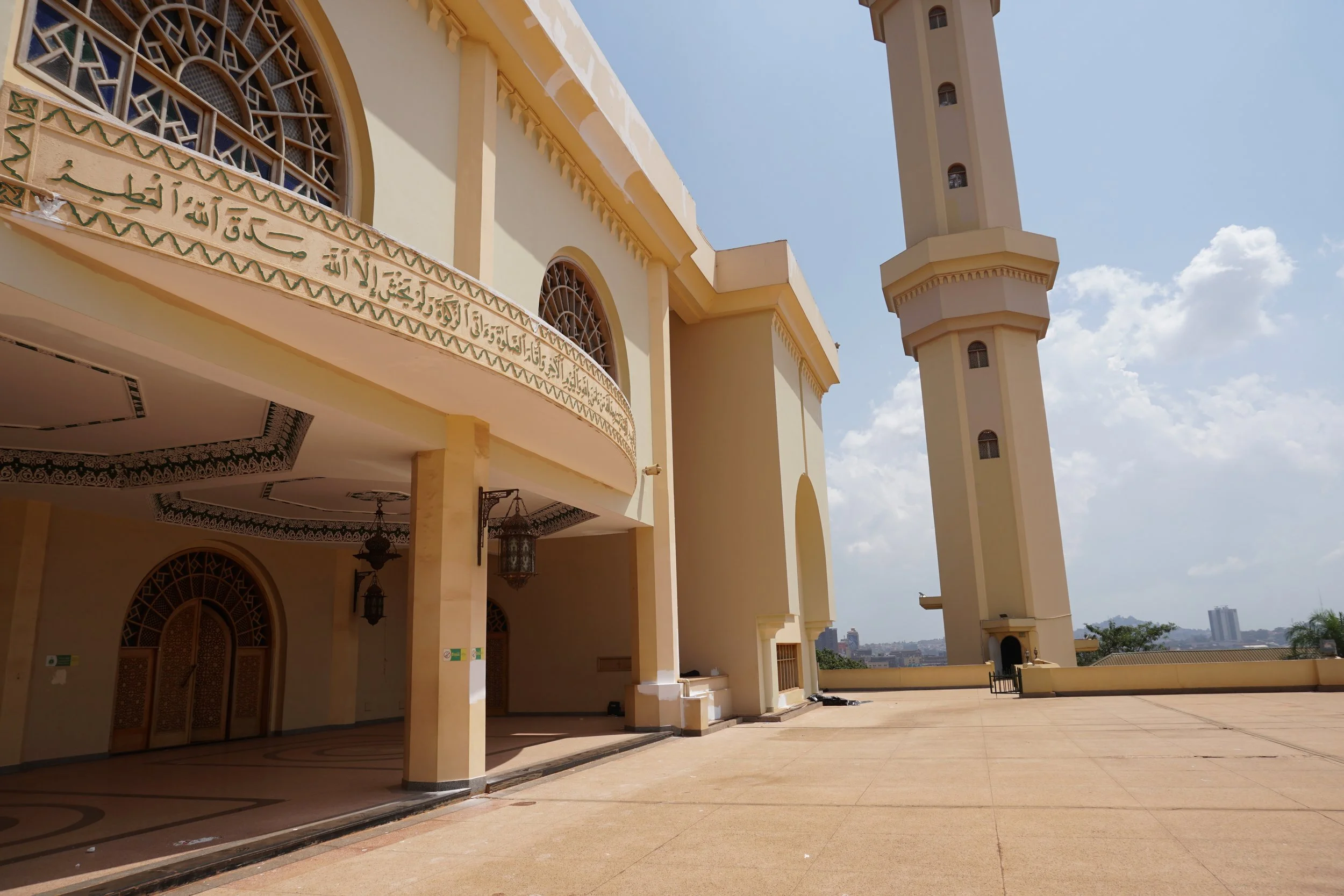 Exterior view of a mosque with a tall minaret, arched windows, decorative Islamic calligraphy, and an open terrace under a blue sky with clouds.