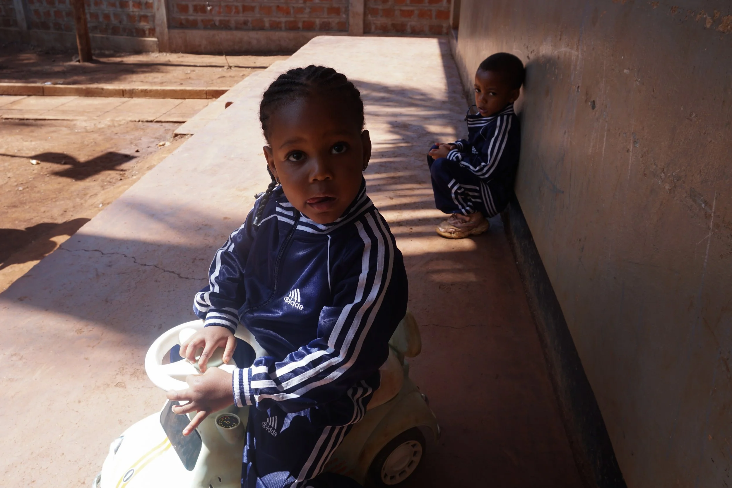 Two young children wearing matching navy blue Adidas track suits sitting outside; one child on a small toy car and the other leaning against a wall