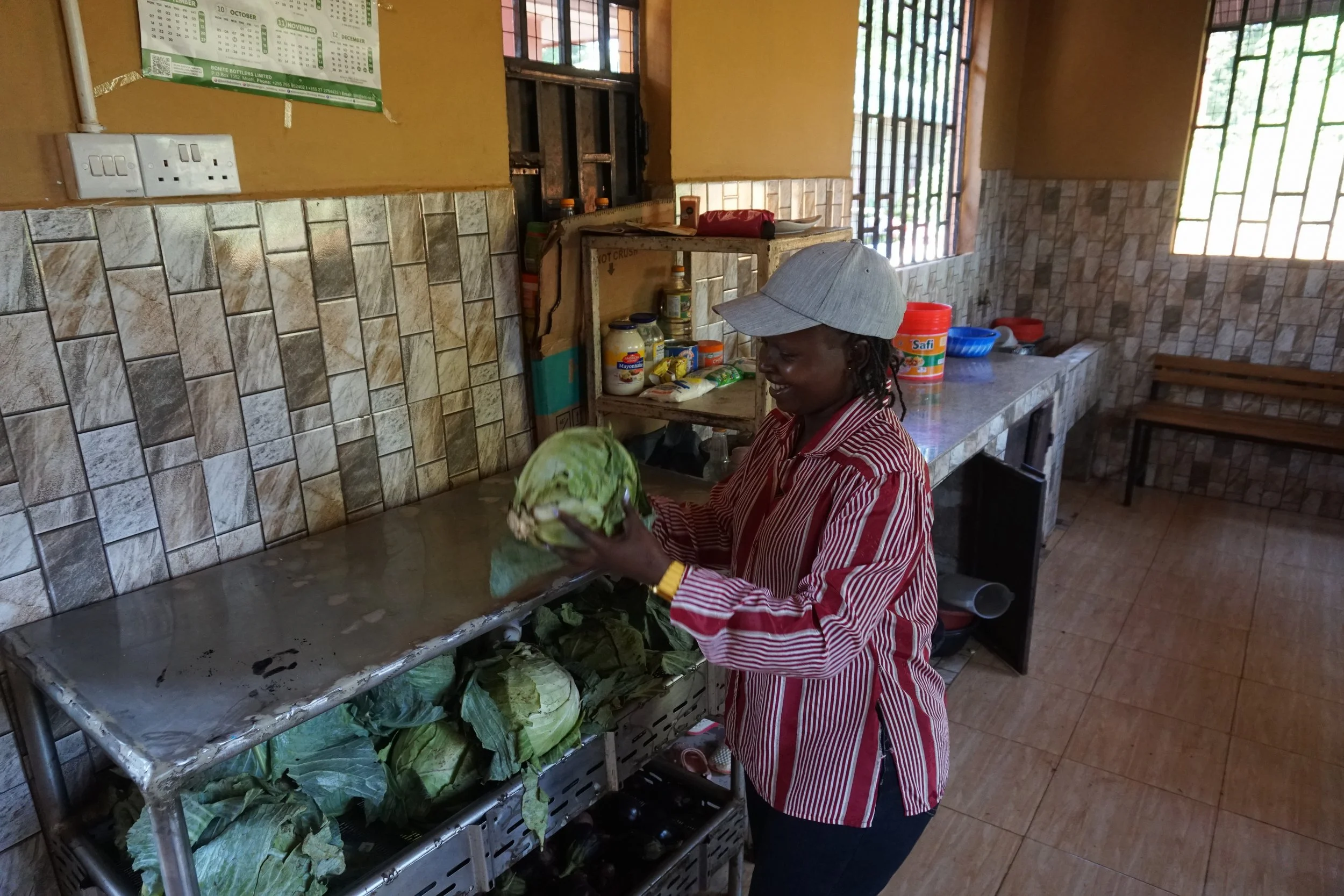 A woman wearing a striped red and white shirt and a gray baseball cap is holding a cabbage in a kitchen with tiled walls and counter.