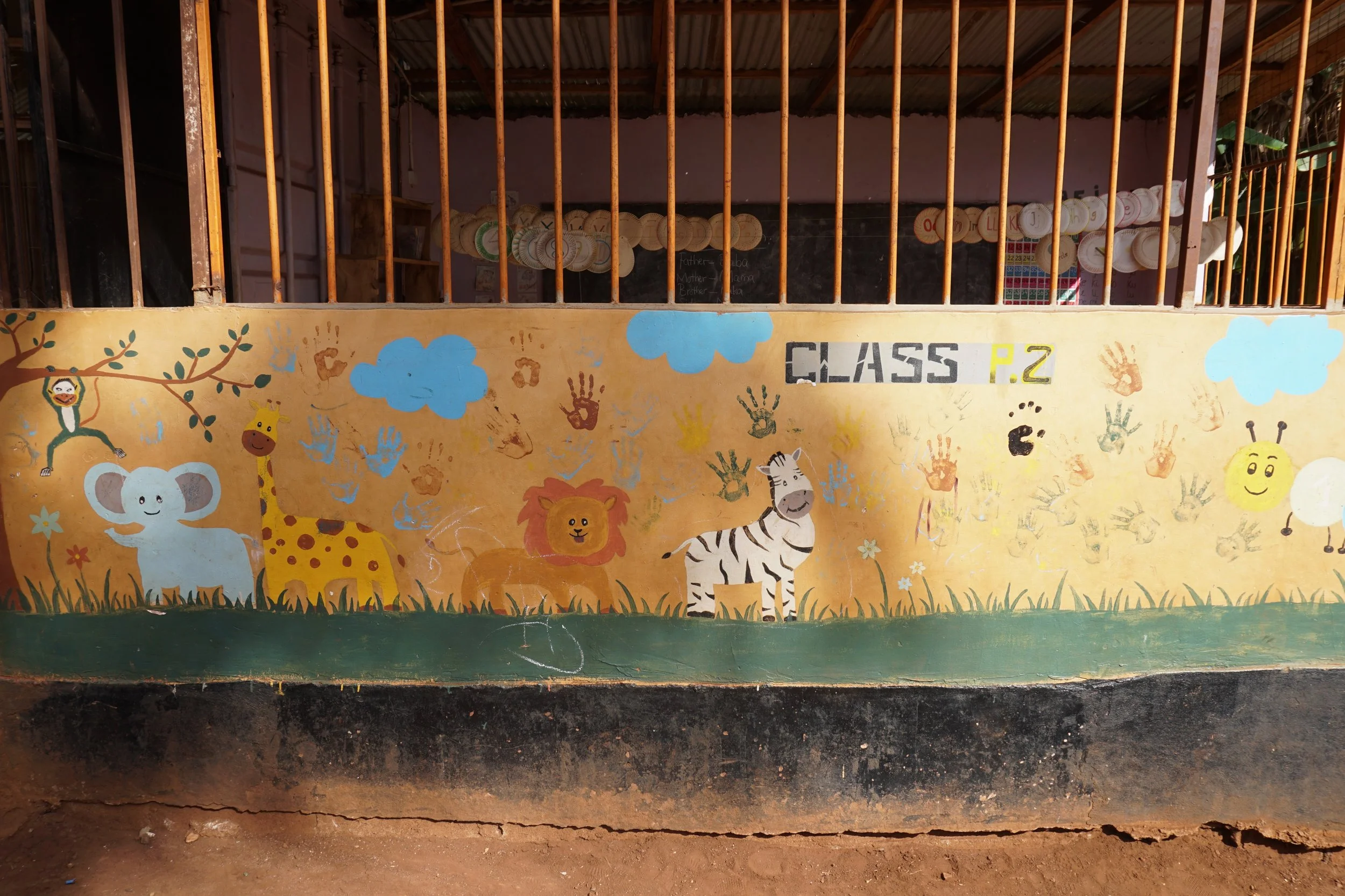 Colorful mural featuring a monkey, giraffe, lion, elephant, and zebra, with handprints and clouds, on a school wall labeled 'Class P.2'.