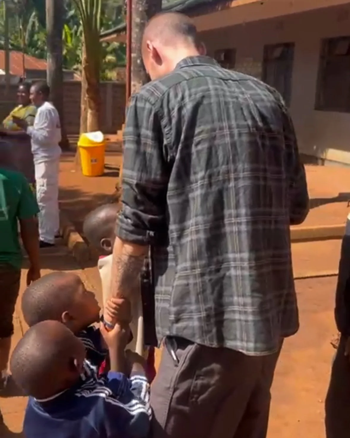 A man with tattoos on his arms is holding hands with a young child in an outdoor setting. Several children and adults are visible in the background, with some standing and others walking on a dirt ground. The scene appears to be in a rural or semi-ur