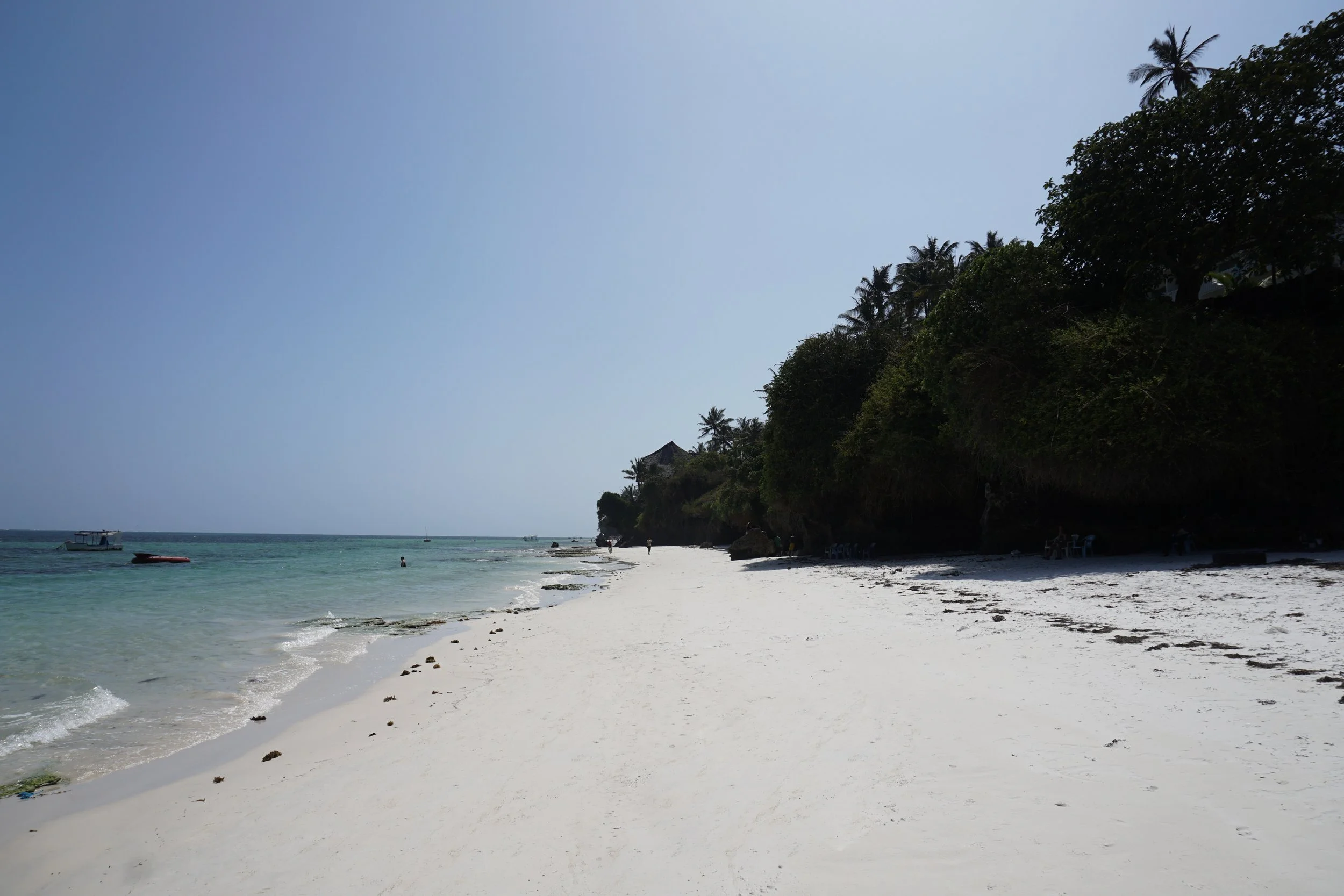 Sunny tropical beach with white sand, clear water, boats, and lush green trees on the right.