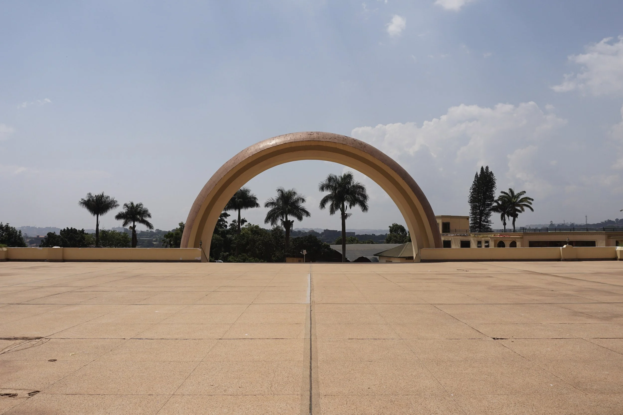 An open paved area with a large beige and pink archway in the background, trees and a partly cloudy sky visible beyond.