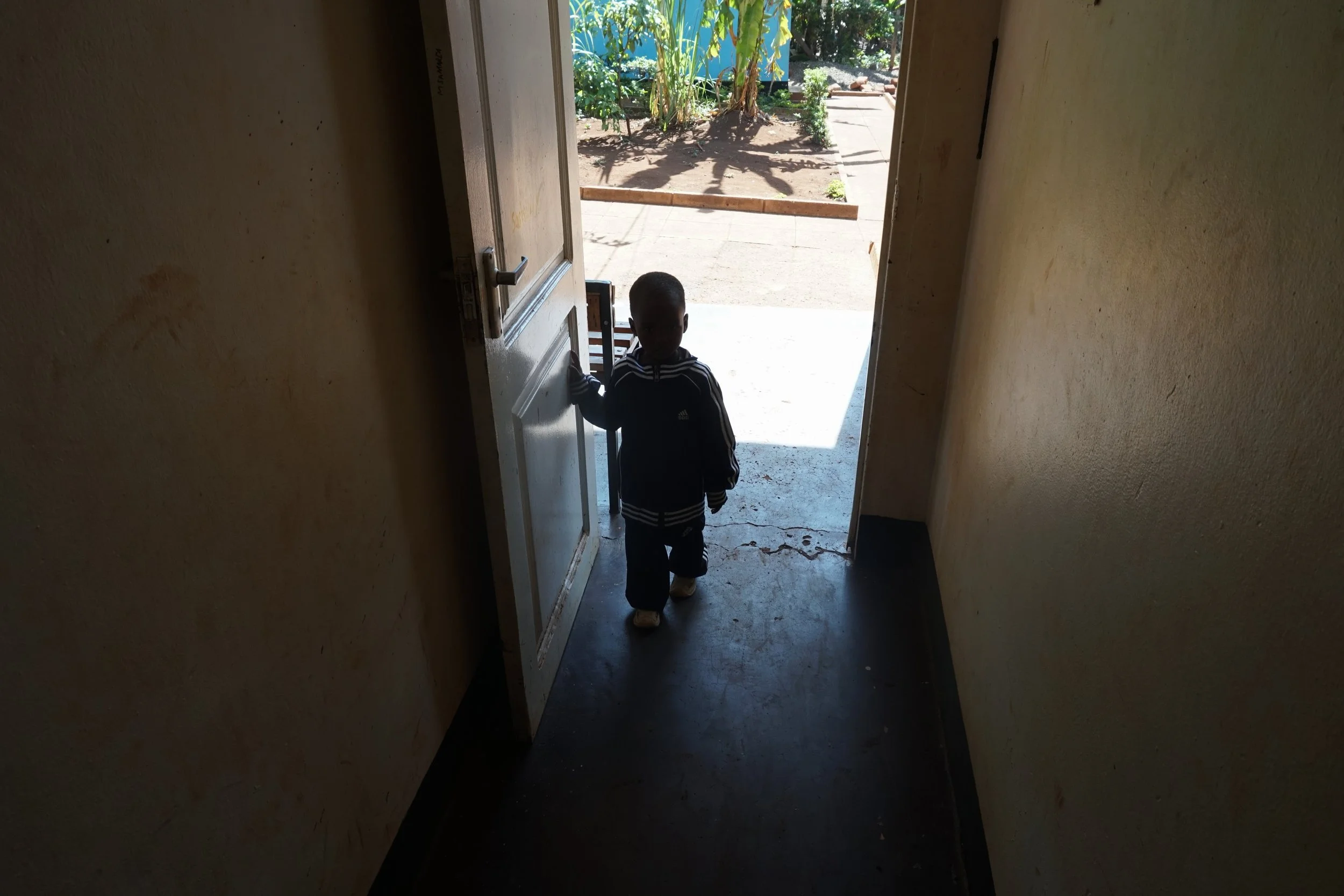 A young boy standing at the doorway inside a room, with sunlight illuminating the area outside, revealing some plants and a dirt ground.