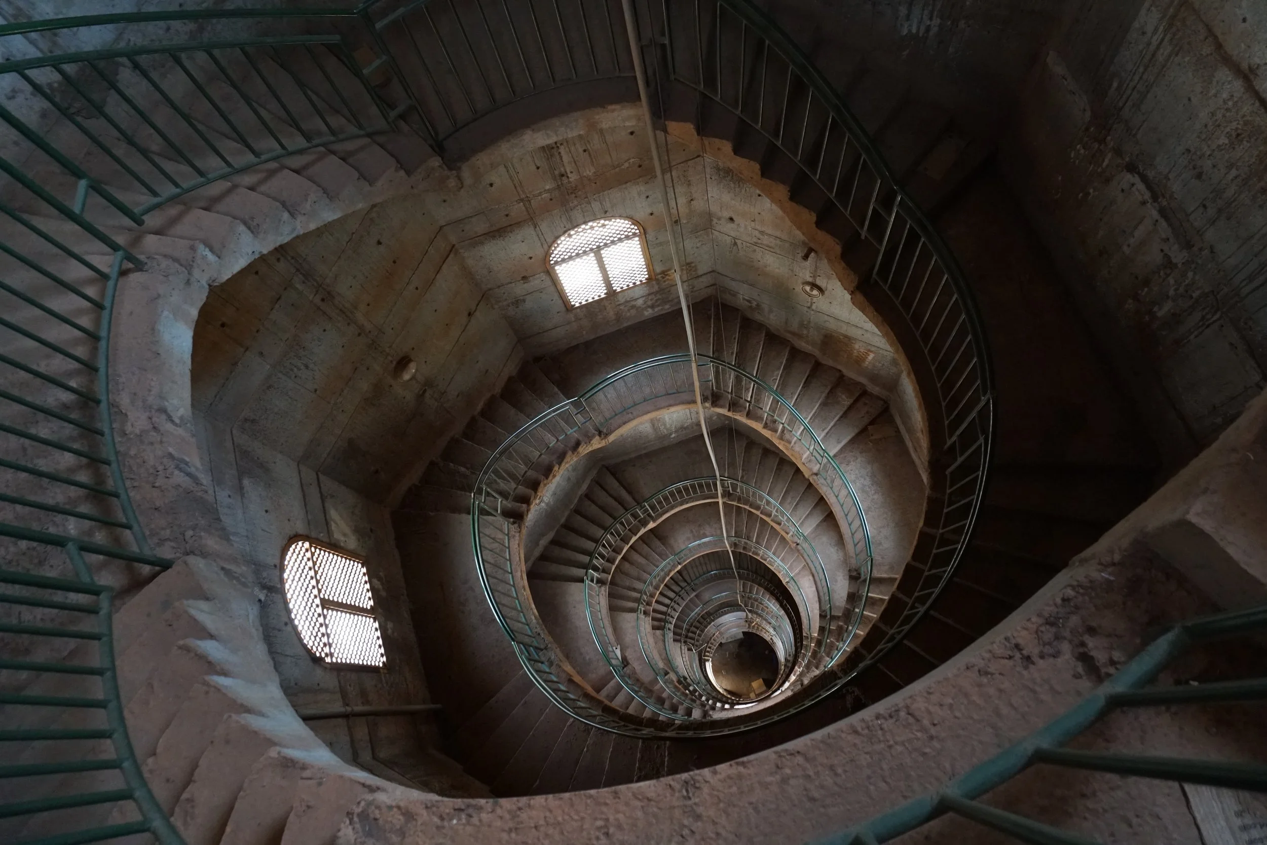 View from the top of a tall spiral staircase looking down into the center, with concrete walls and metal railings.