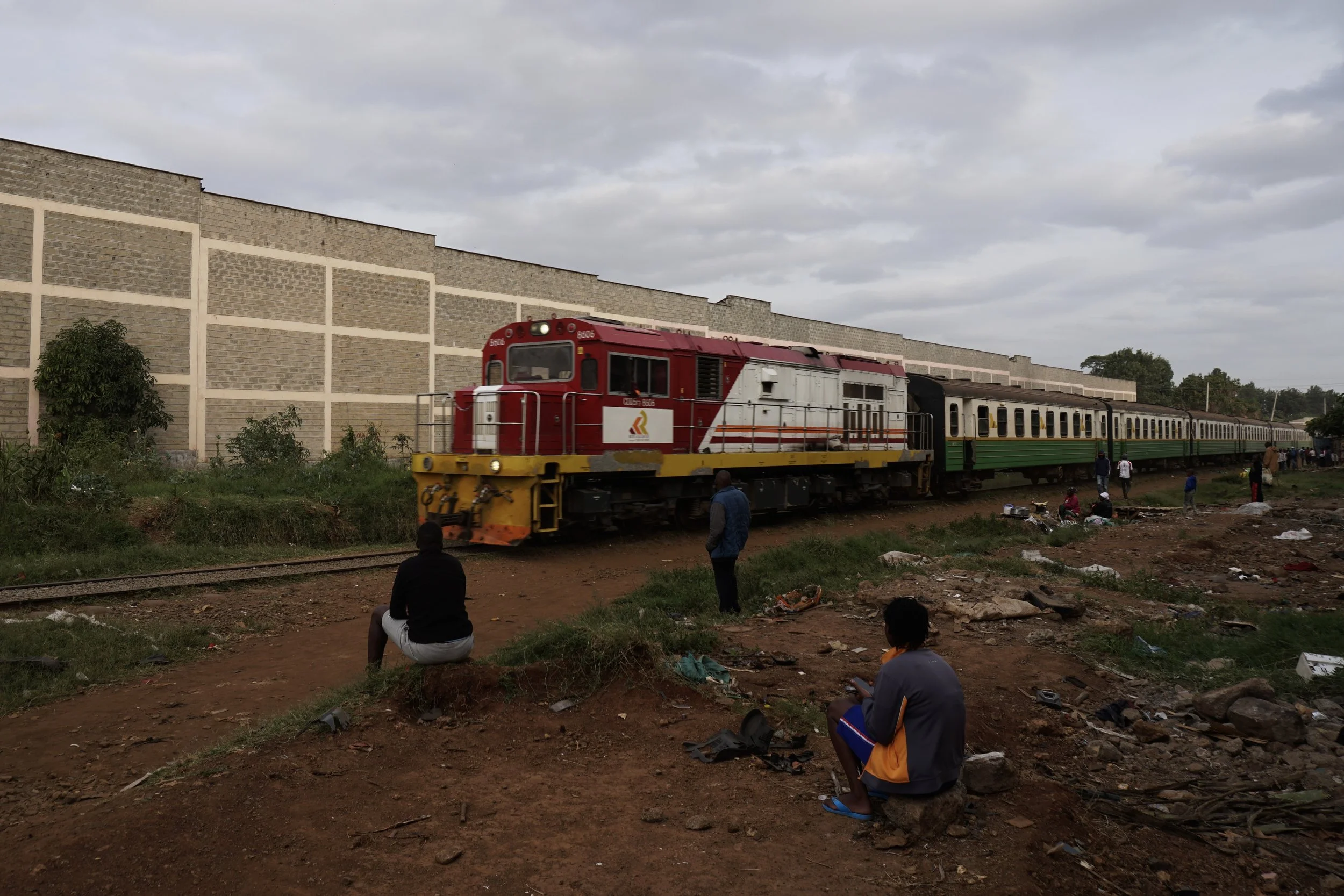 A red and yellow train passes by a group of people sitting and standing along a dirt path next to the railway tracks, with a large concrete wall and cloudy sky in the background.