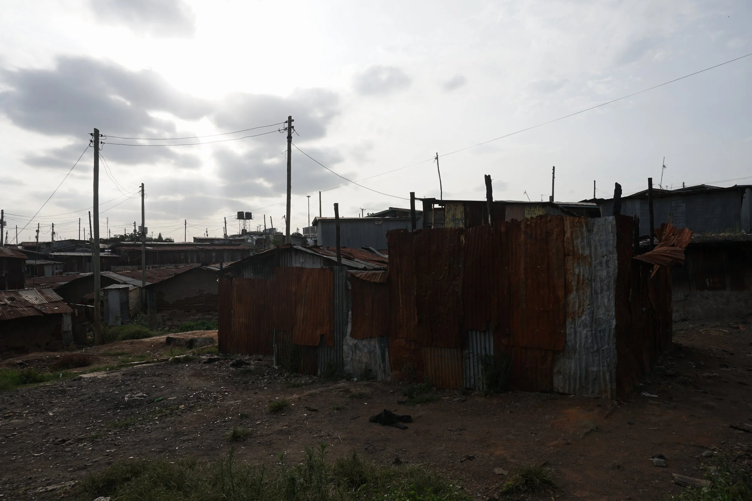A shantytown with makeshift houses made of corrugated metal sheets in a developing area, with utility poles and overcast sky.