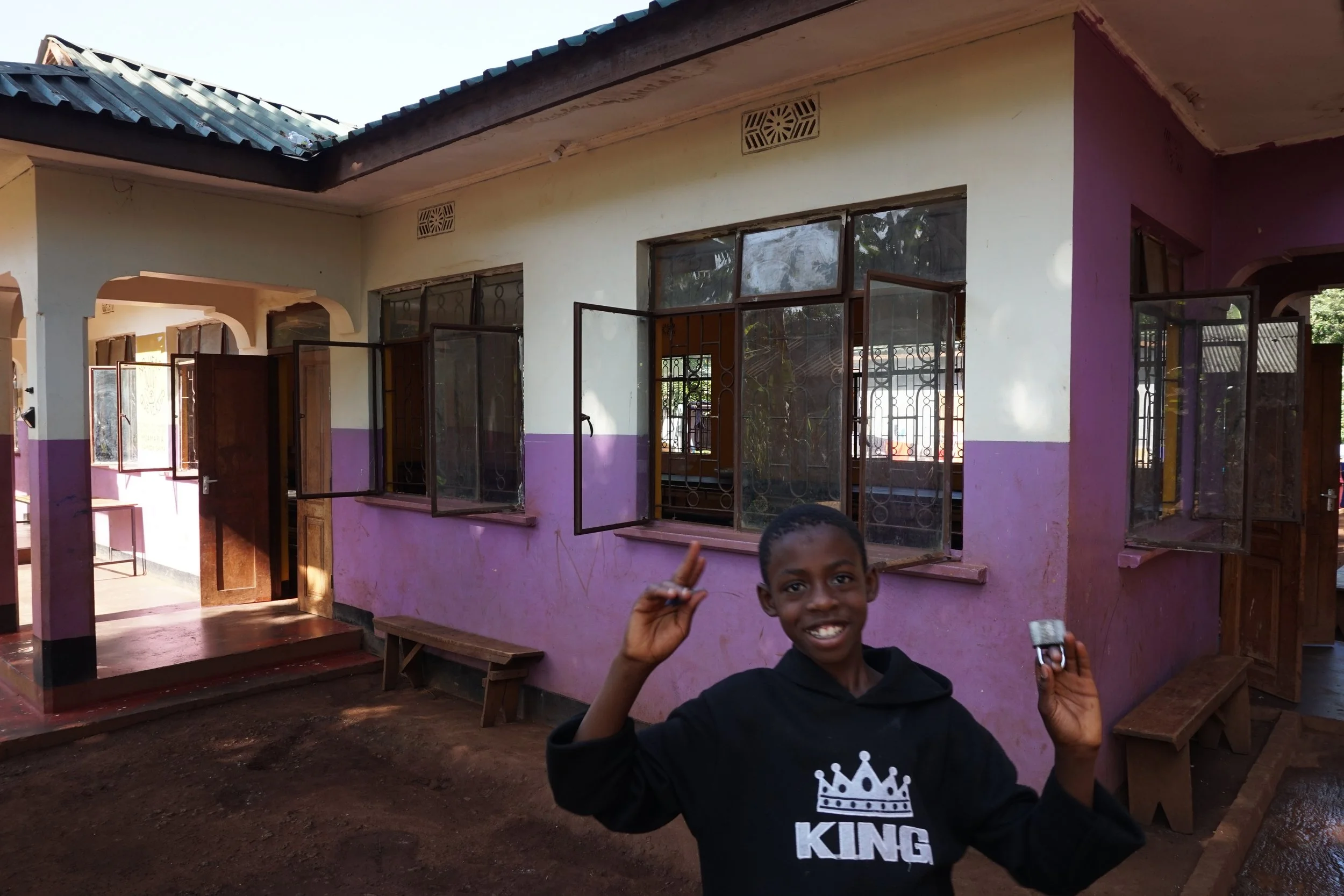 A smiling boy standing outside a colorful classroom building, holding a small object in one hand and making a peace sign with the other.