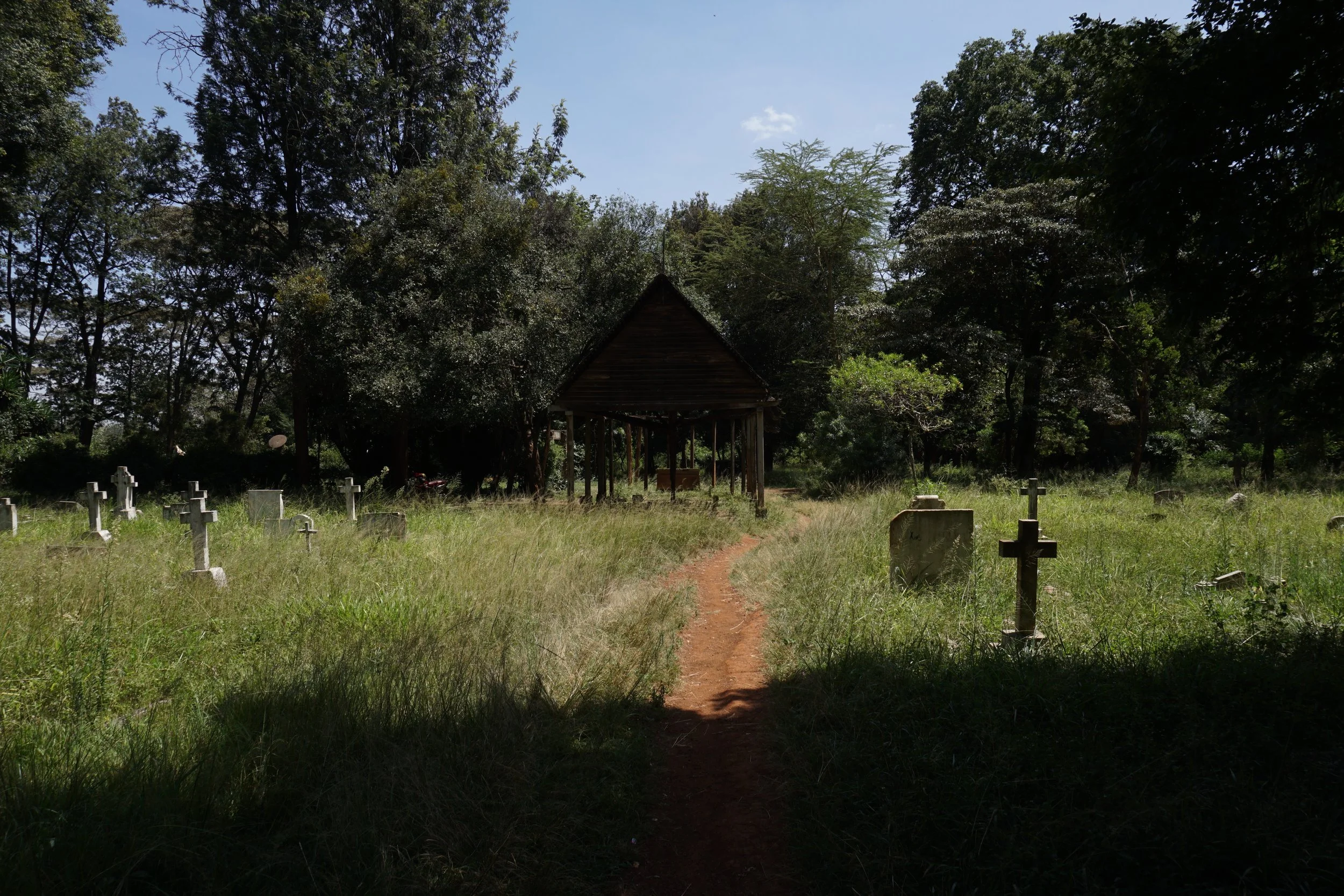 A small dirt path leads through a grassy cemetery with stone crosses and headstones, ending at a wooden shelter, surrounded by trees under a clear blue sky.
