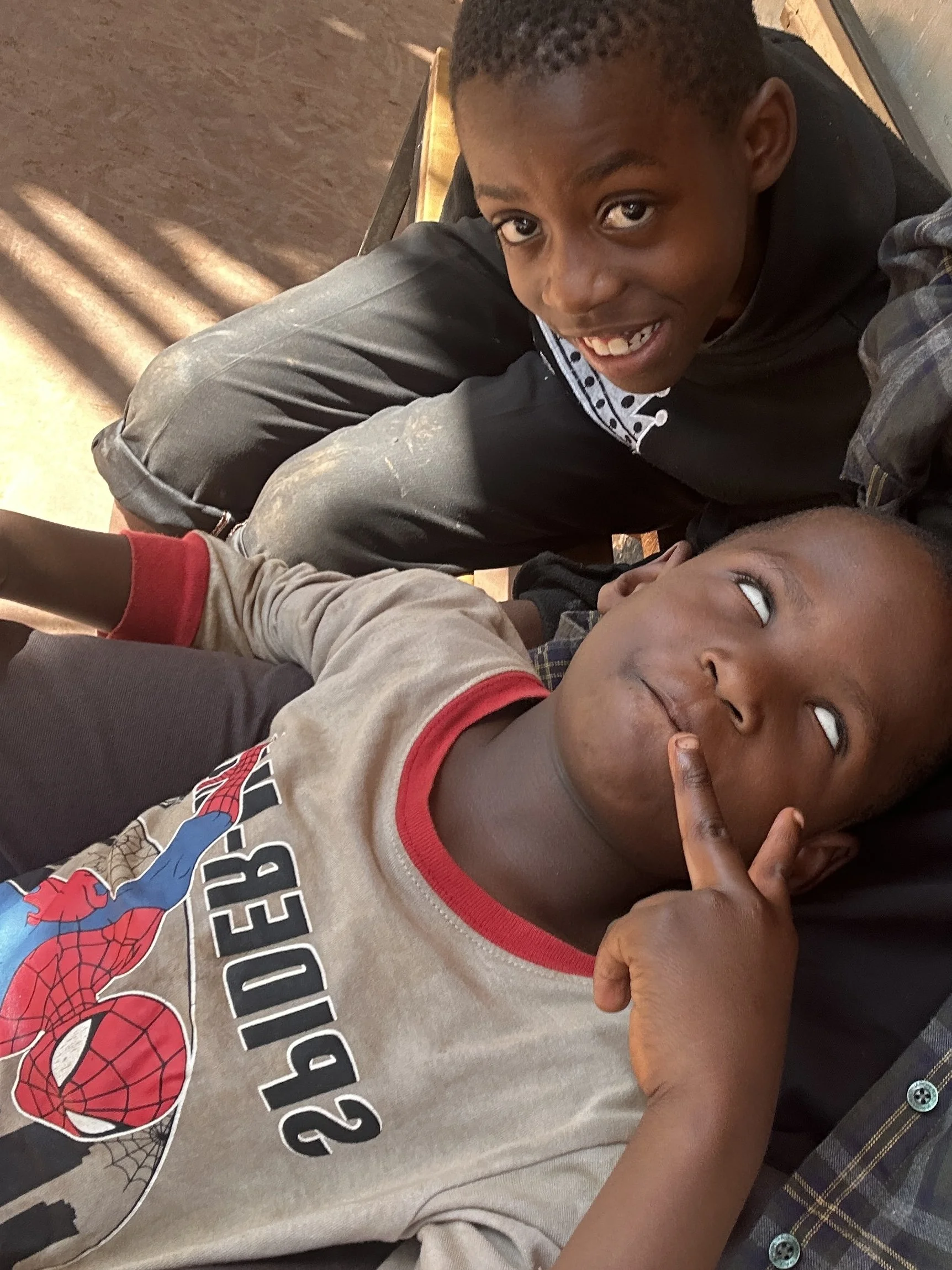 Two young boys lying on a bed indoors, one with a big smile and looking up, the other with a thoughtful expression and finger on lips, wearing a Spider-Man shirt.