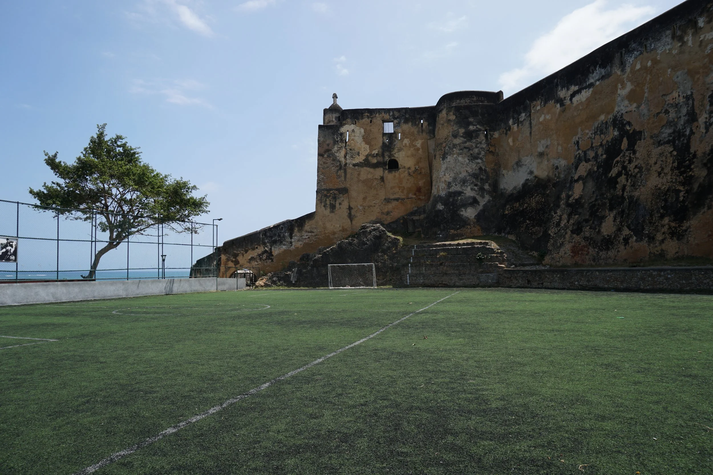 Empty soccer field with goalpost, large tree, stone fortress wall in the background, and blue sky.