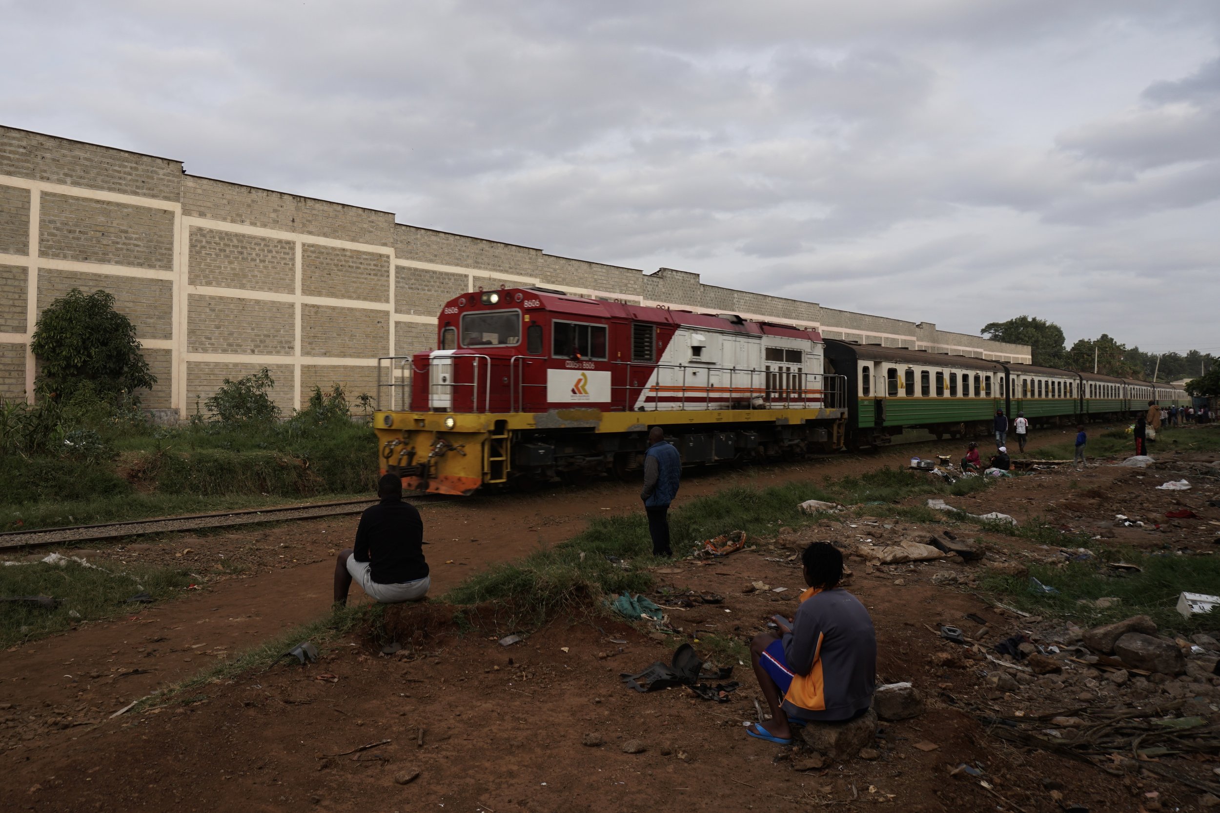 A red train moving along a railway track with passengers sitting nearby in an outdoor setting, some on rocks and others on the ground, with a large brick wall in the background under a cloudy sky.