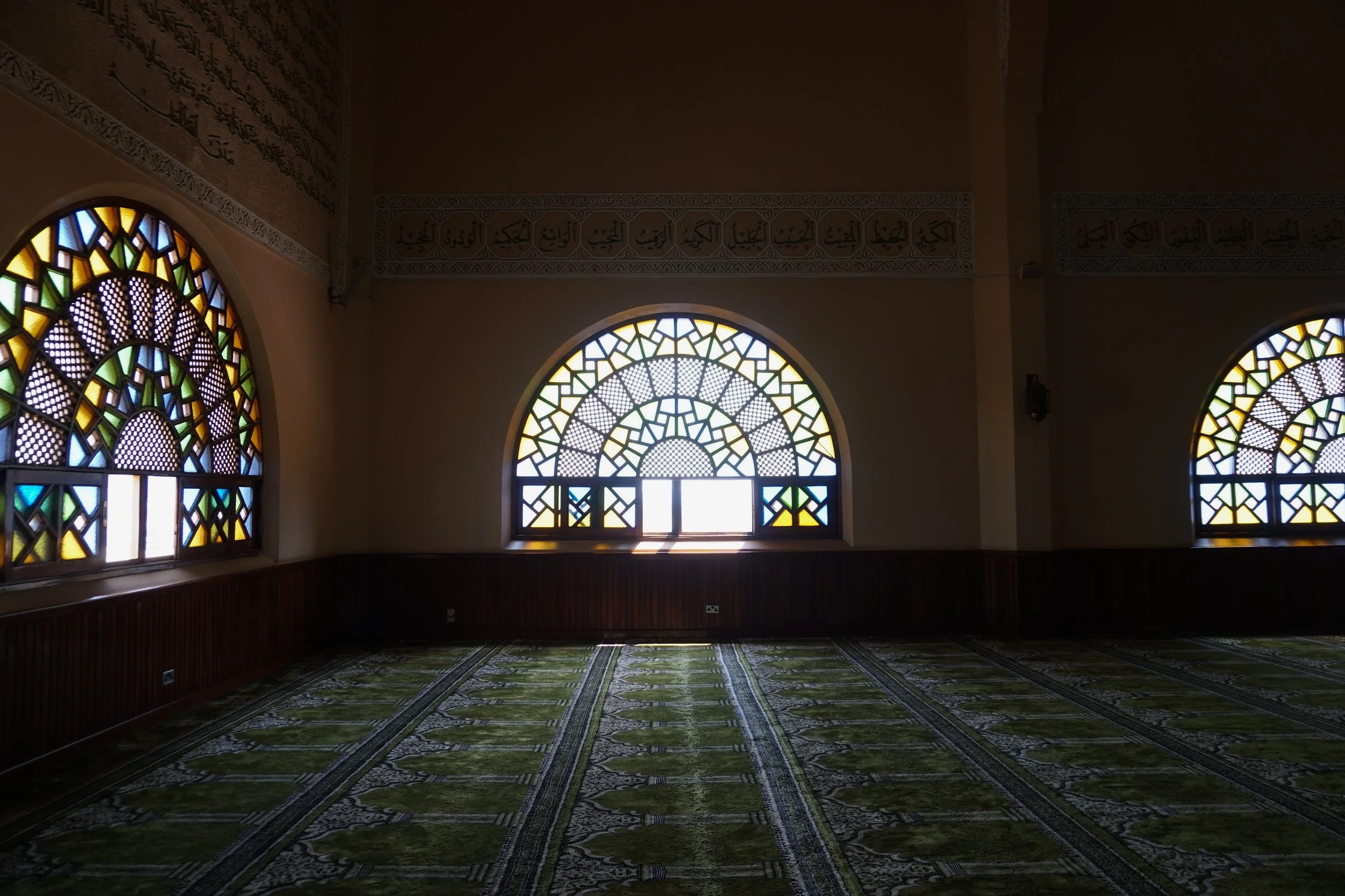 Interior of a mosque with arched stained glass windows and patterned green carpet.