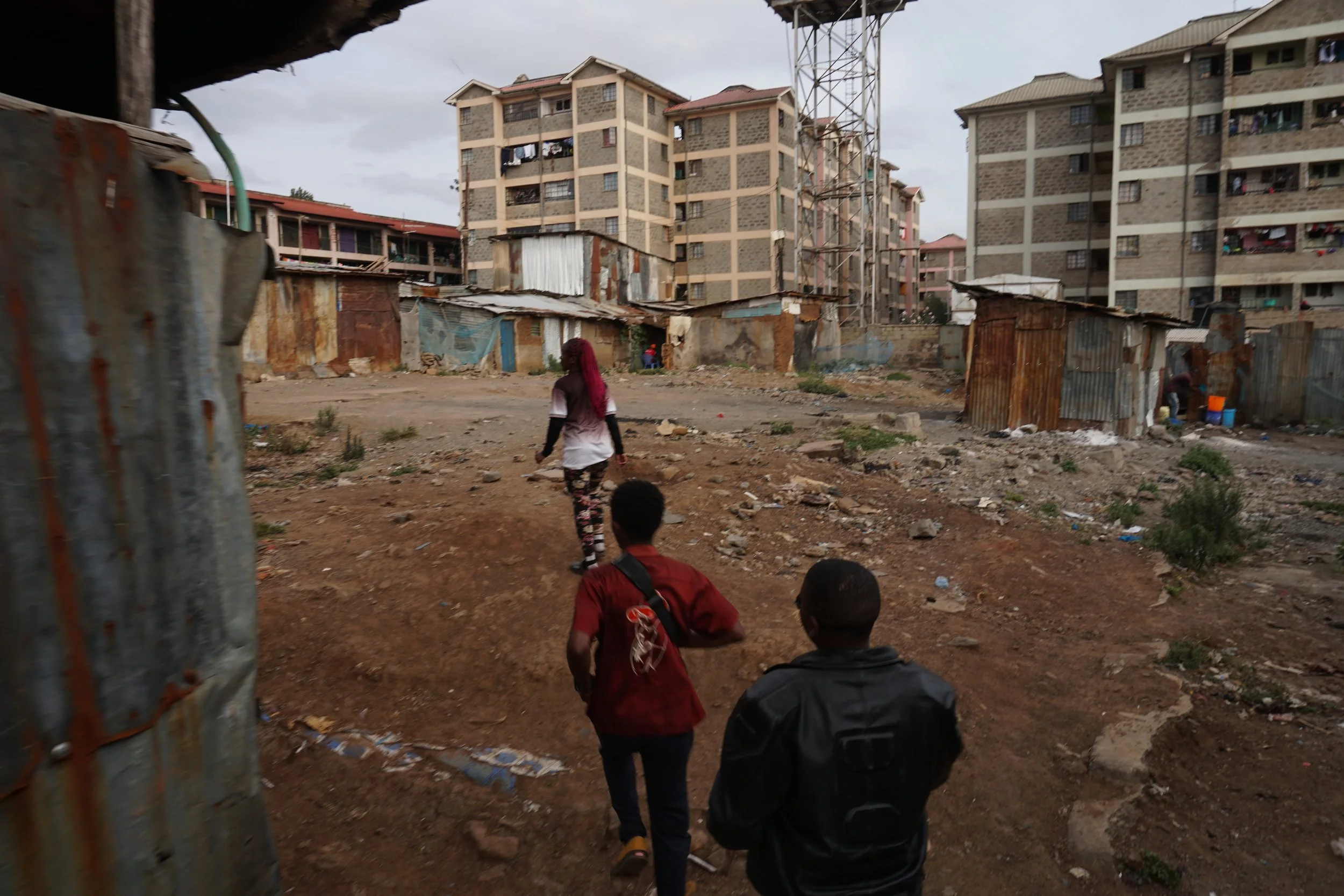 Three people walking through a dirt area in front of makeshift metal shacks with high-rise buildings in the background.