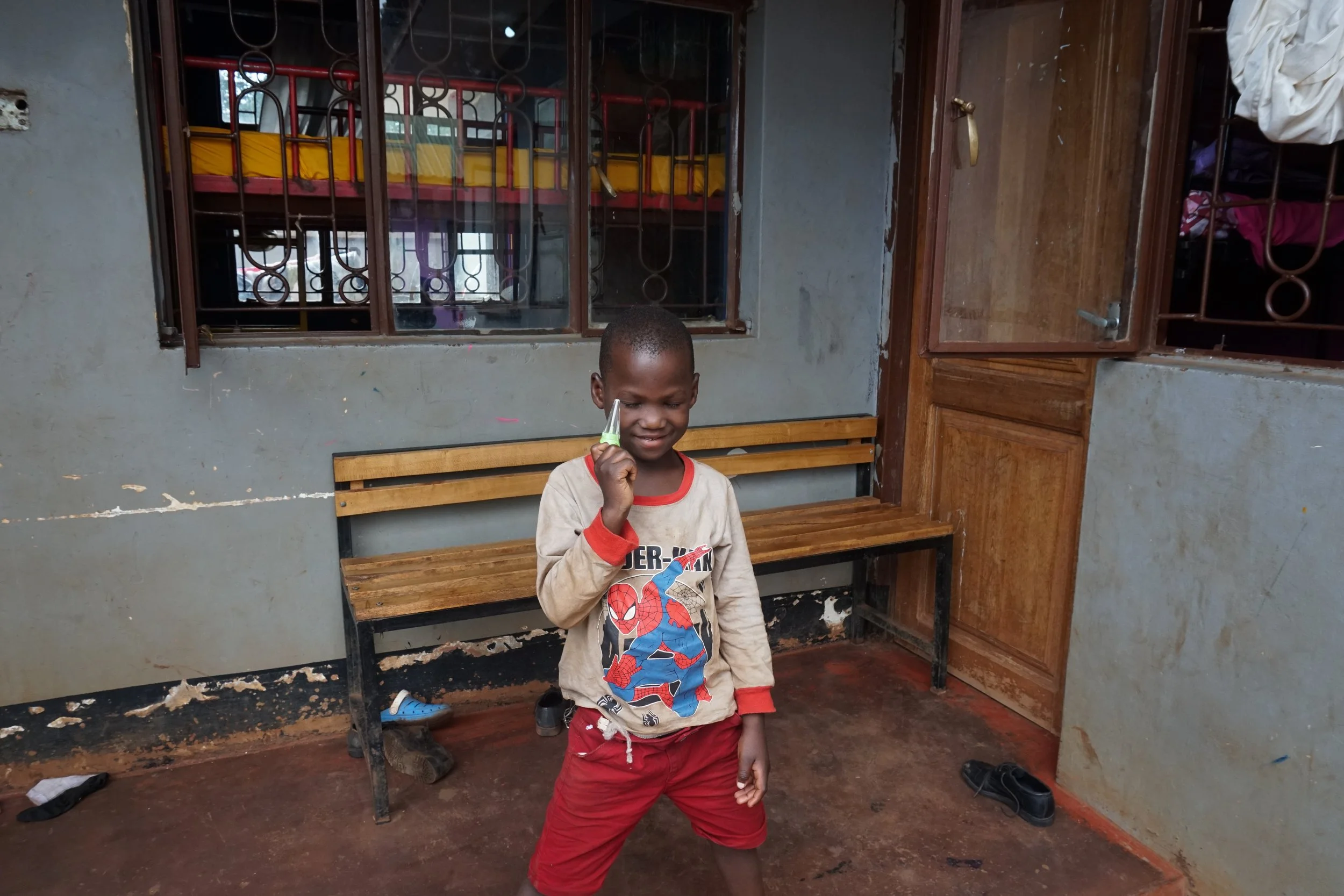 Young boy holding a small toy rocket in a room with a wooden bench, shoes on the floor, and a window with metal bars.