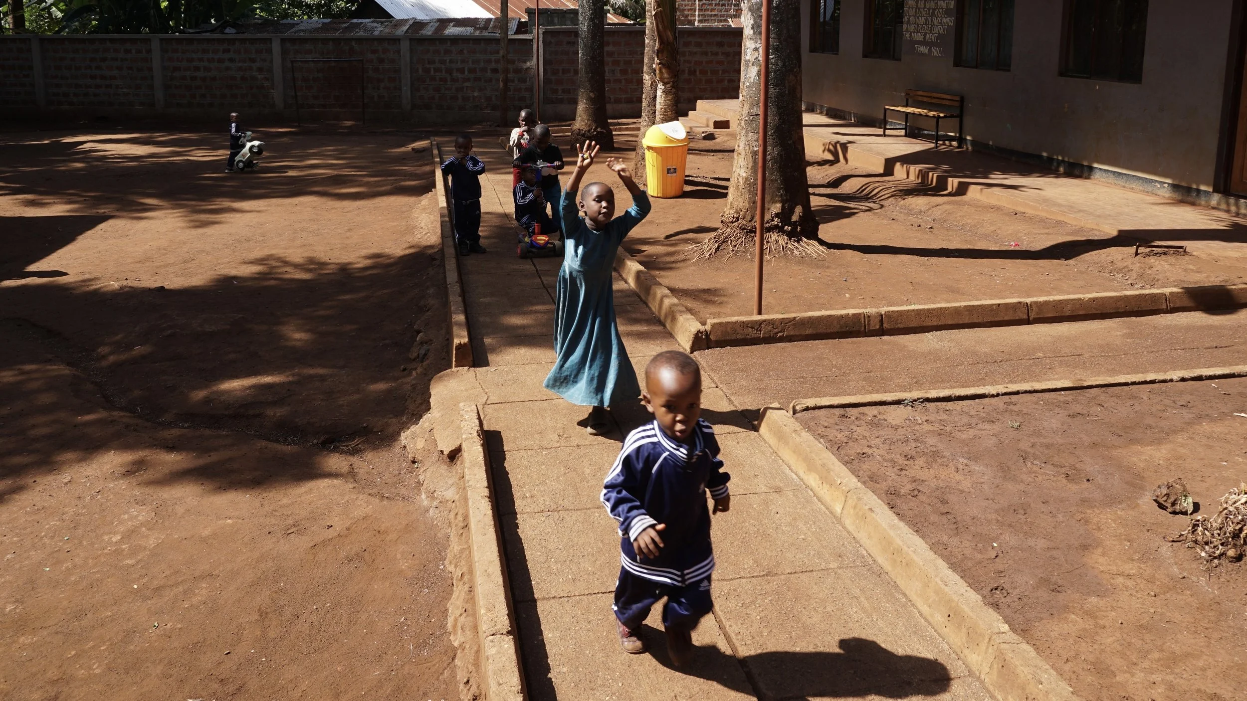 Young children playing outside on a dirt playground, some are on scooters and others are walking. Two boys are in the foreground, one looking at the camera and the other raising his hand. There are trees, a yellow trash can, and a building with a ben